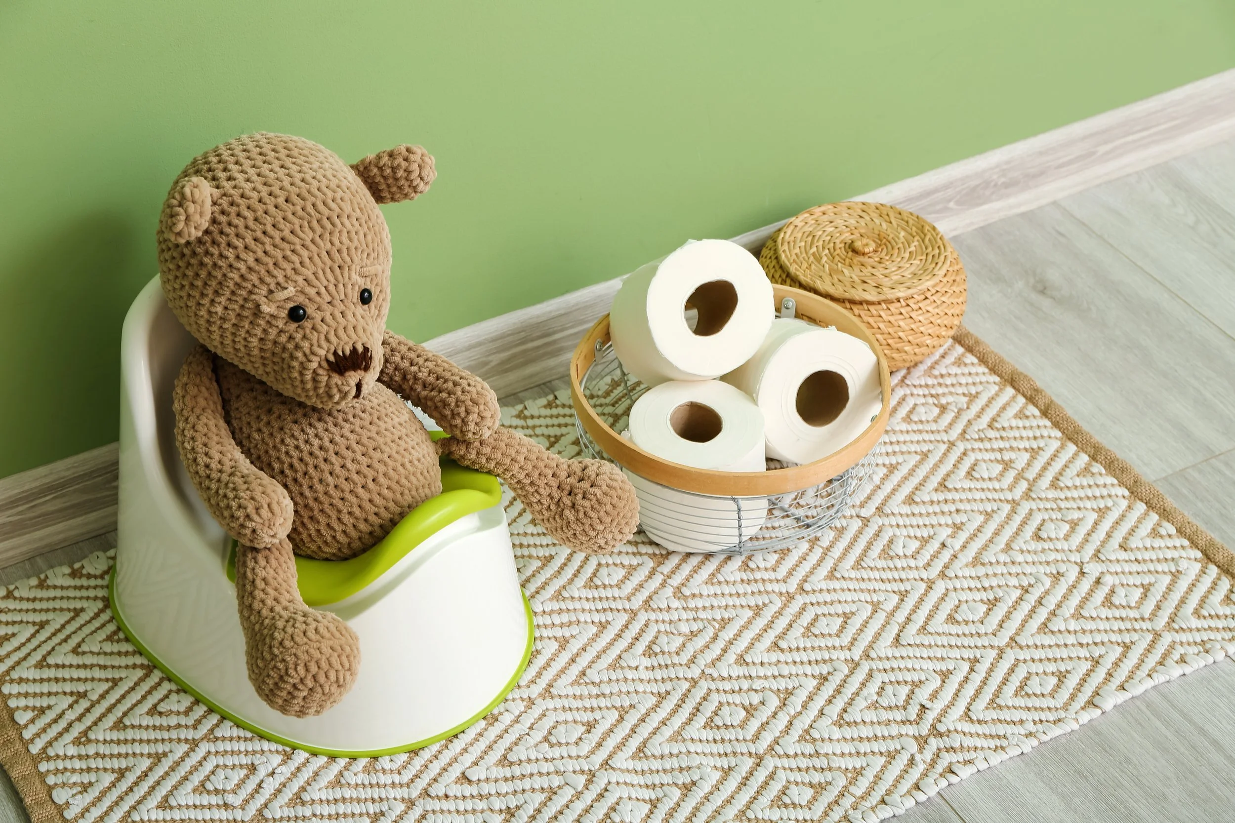 A crocheted teddy bear sitting in a child's potty, next to a basket of toilet paper rolls and a woven container, on a decorative rug against a green wall.