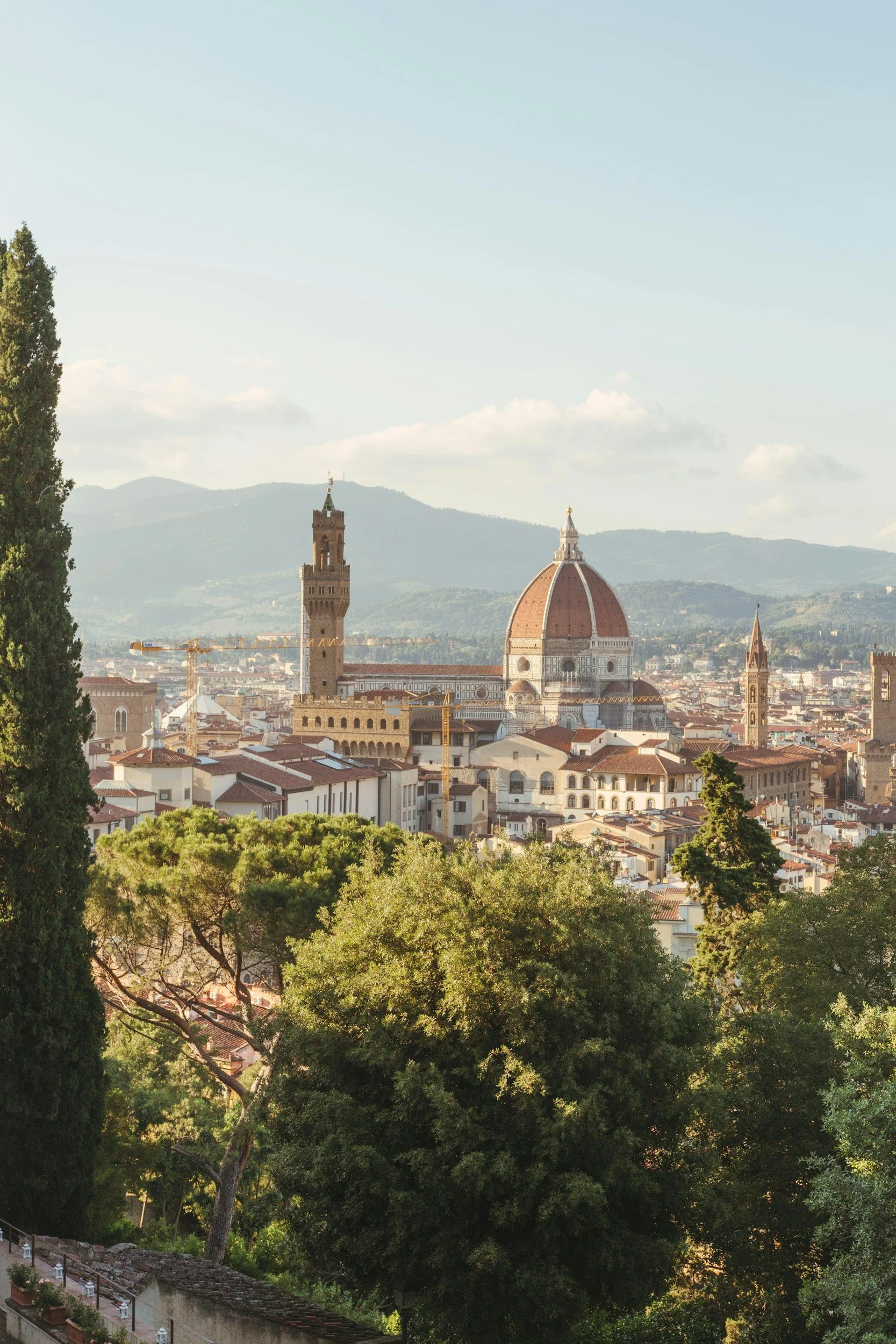 View of Florence, Italy, with the Florence Cathedral and Giotto's campanile in the cityscape, framed by lush green trees in the foreground.