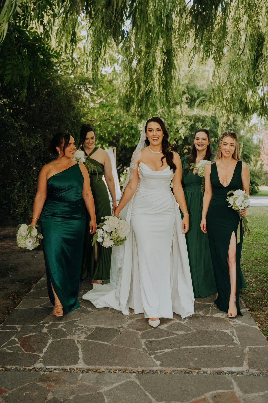 Bride in white wedding dress walking with five bridesmaids in green dresses, holding bouquets, under a leafy tree on a stone pathway.