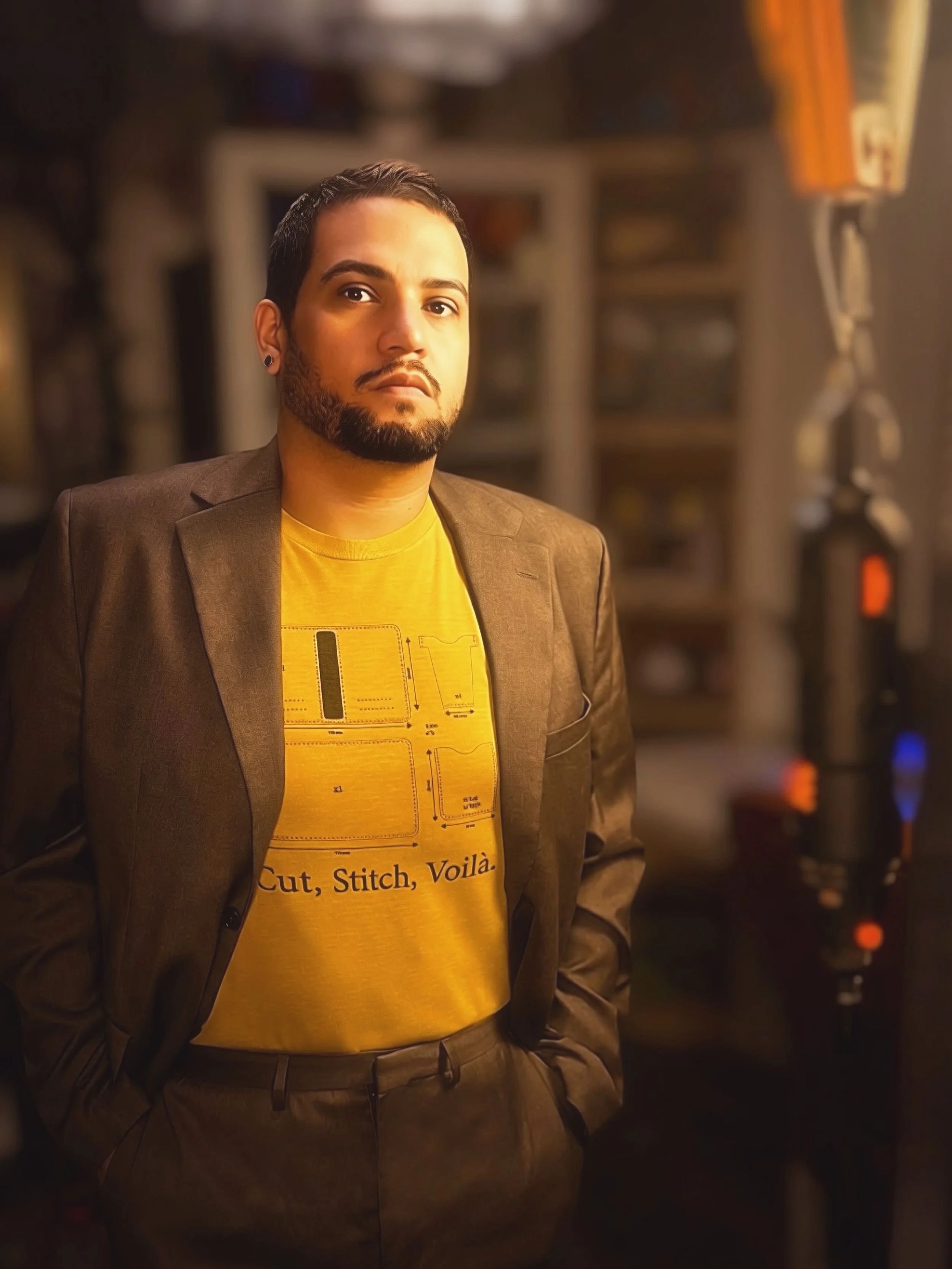 A man with dark hair, beard, and earrings wearing a yellow t-shirt and brown blazer, standing in front of a blurred background with equipment, possibly in a recording studio or radio station.