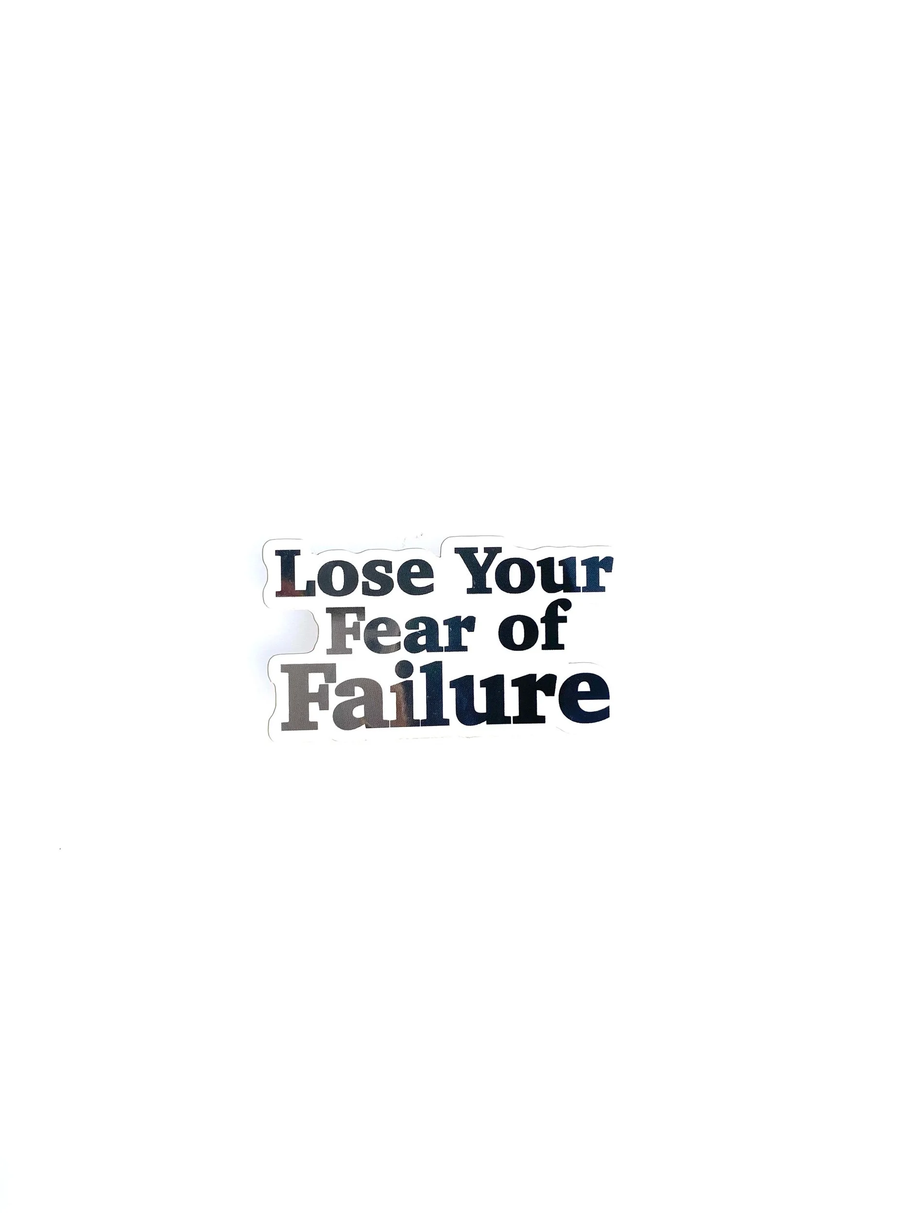 Close-up of a sticker with the words 'Lose Your Fear of Failure' held in front of a black book or box, with a green plant nearby on a white surface.