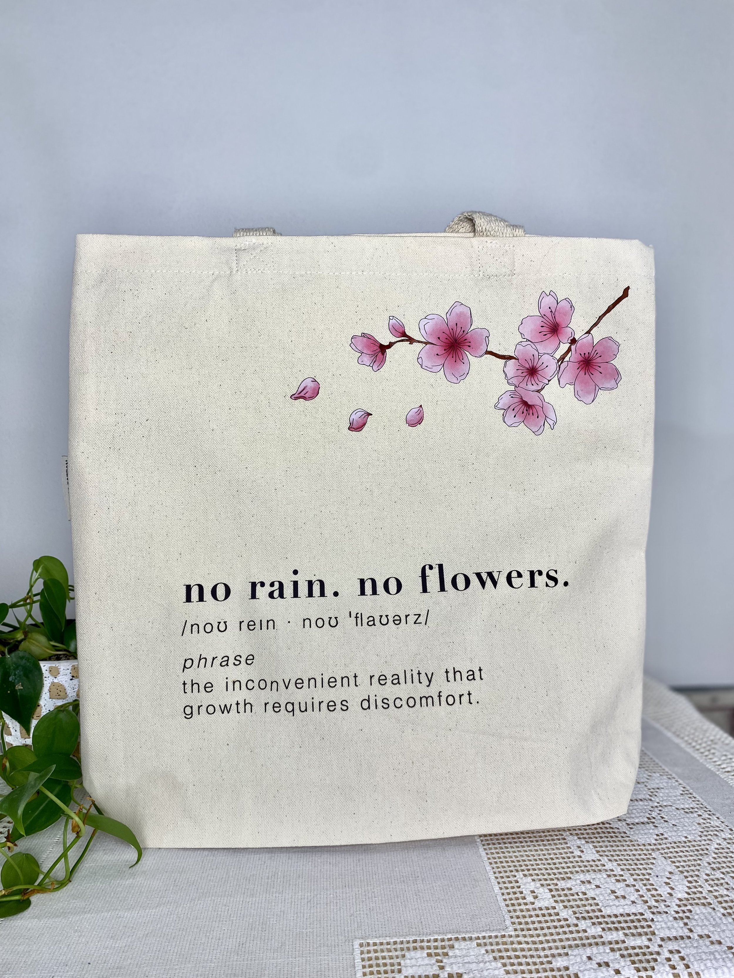 A beige 100% organic cotton tote bag with pink cherry blossom illustrations and the quote 'no rain. no flowers.' written on it.
