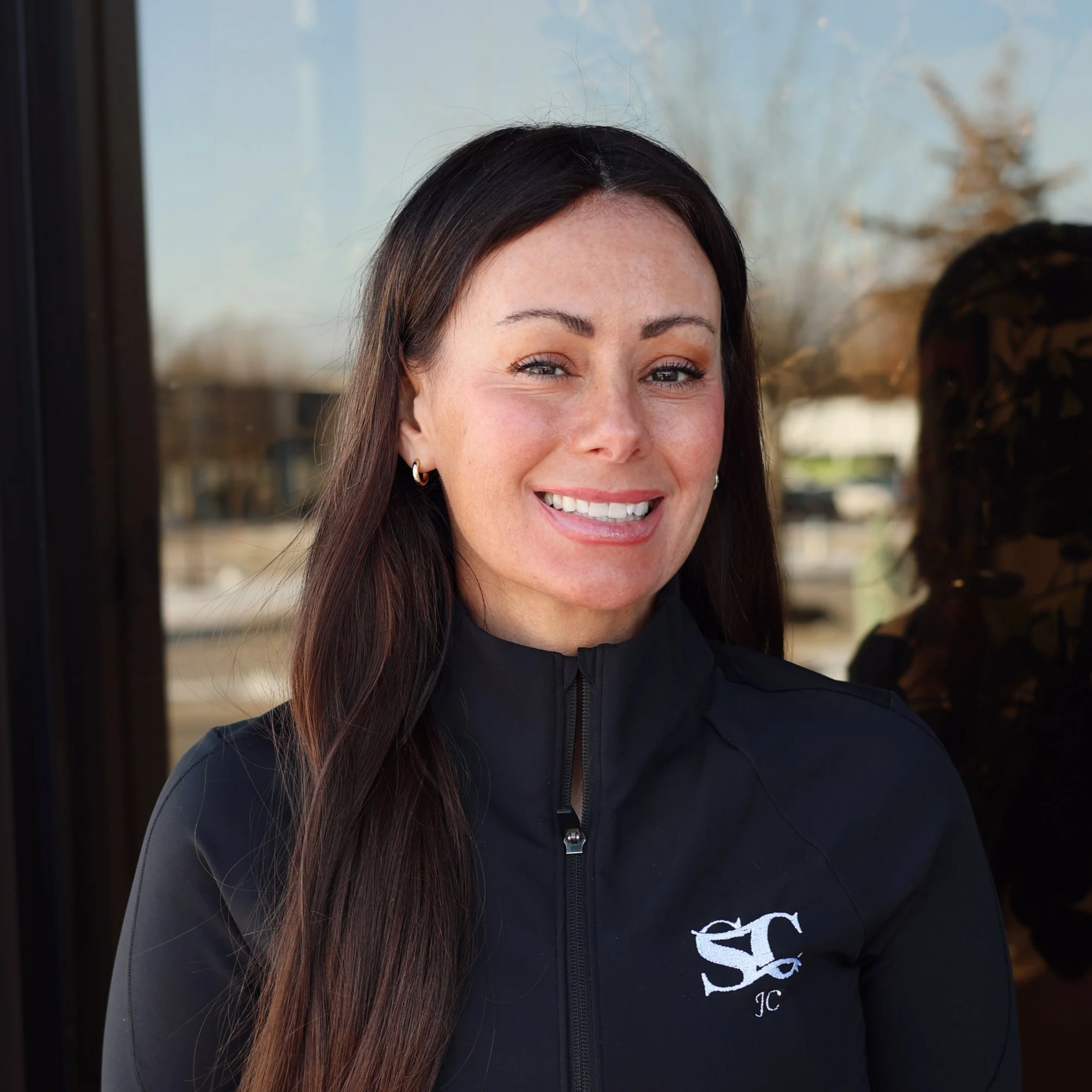 A woman with long, wavy brown hair, wearing a blue jacket and hoop earrings, smiling against a blue background.