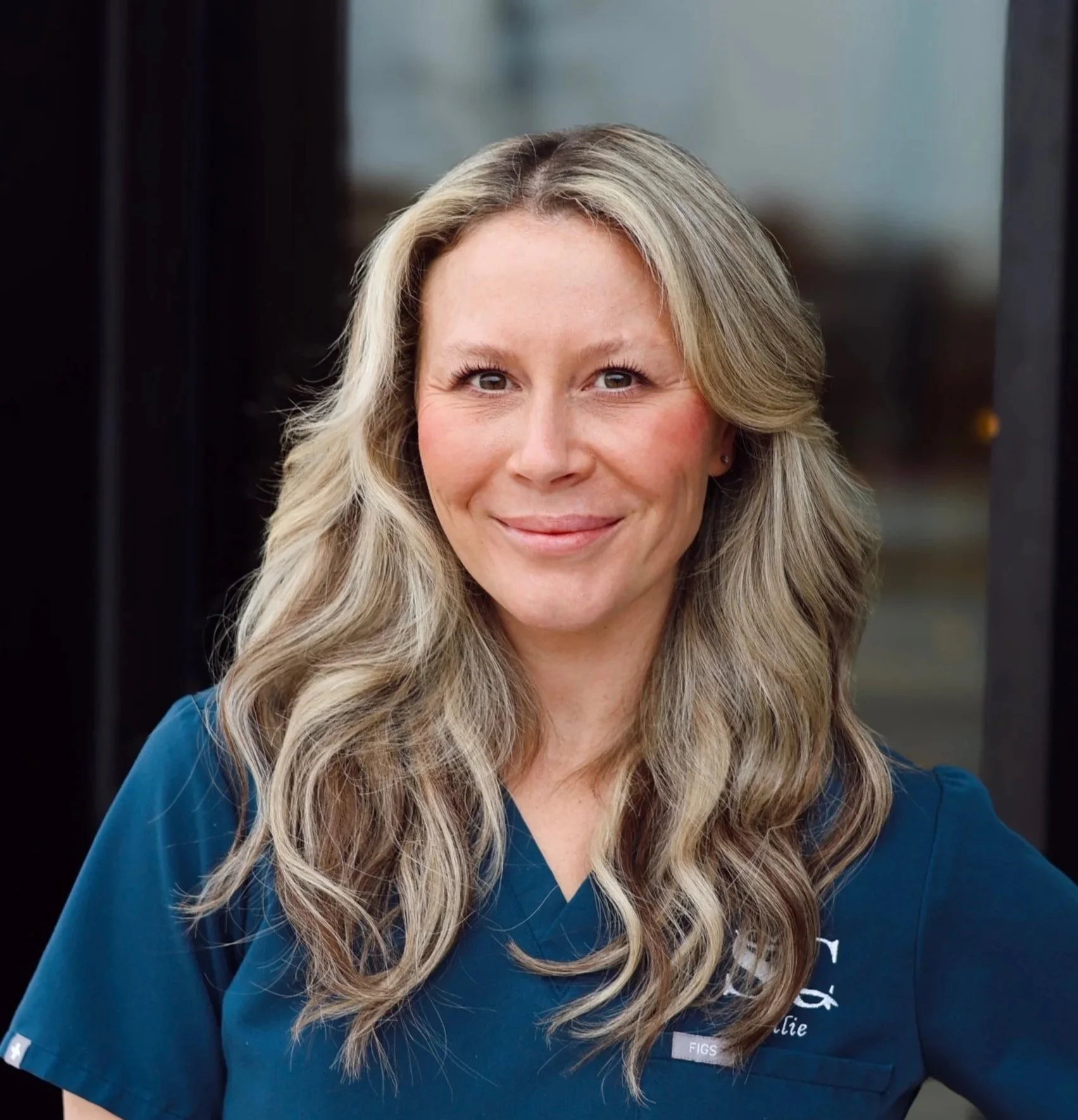 A woman with long wavy blonde hair wearing a blue medical uniform, standing outside near a glass door, smiling at the camera.