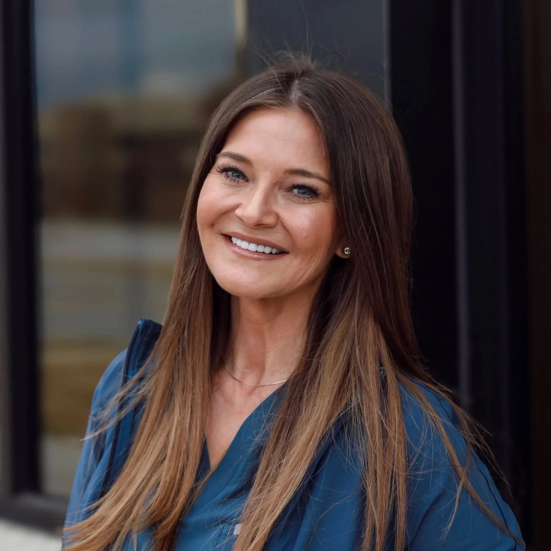 A smiling woman with brown hair and blue eyes wearing a blue jacket, standing outdoors in front of a glass building.