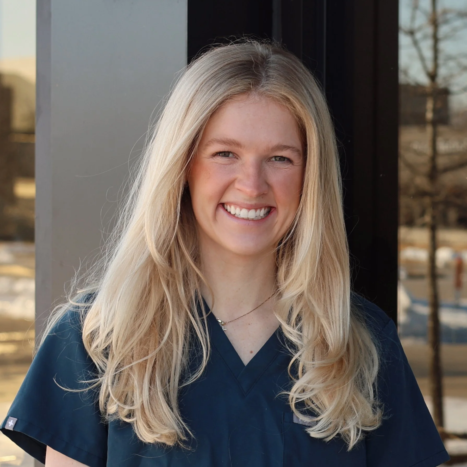 A young woman with long, wavy blonde hair, smiling, standing in front of a glass door.