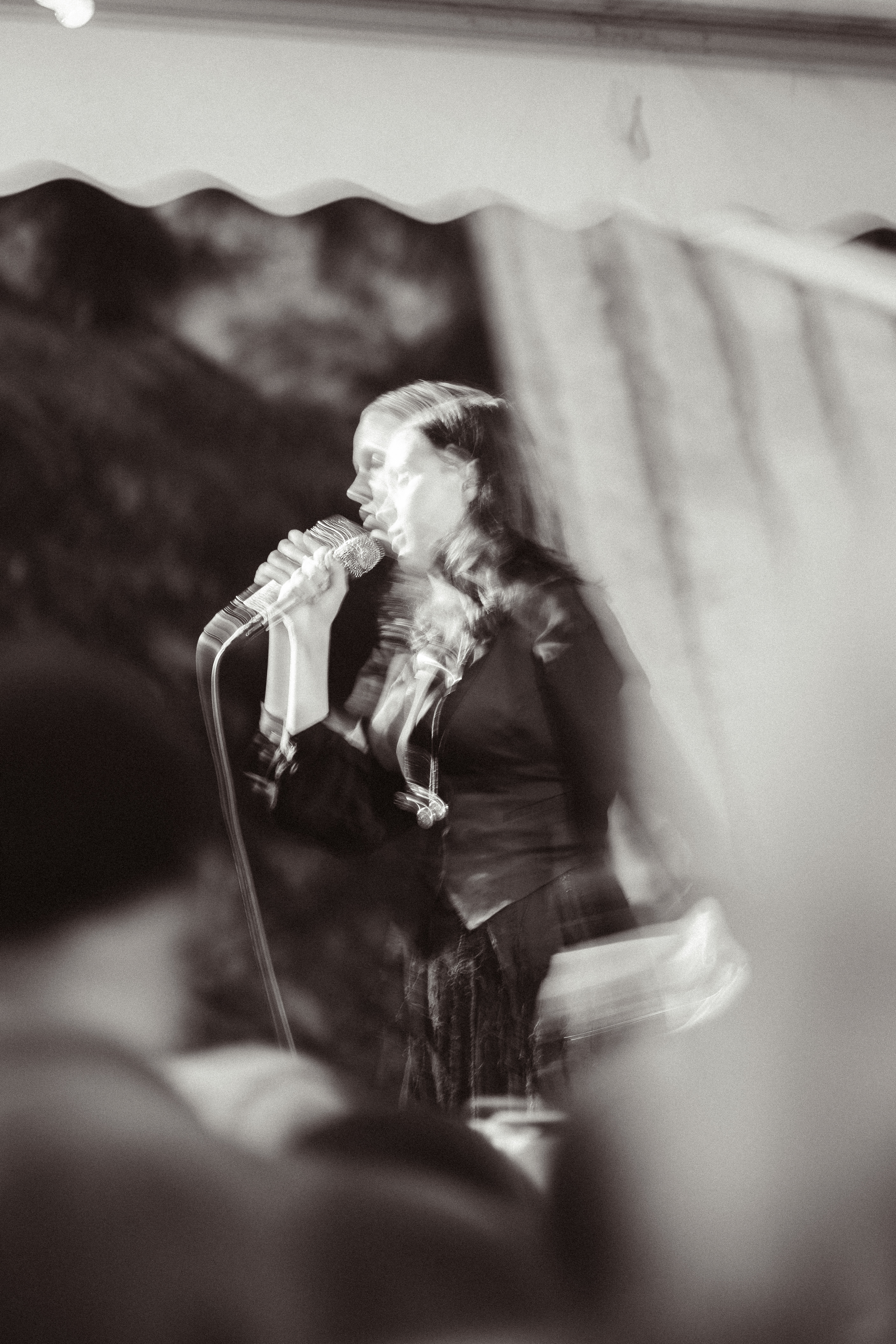 Black and white photo of a woman singing into a microphone, with a ghostly, blurred double exposure effect, possibly during a performance or rehearsal.