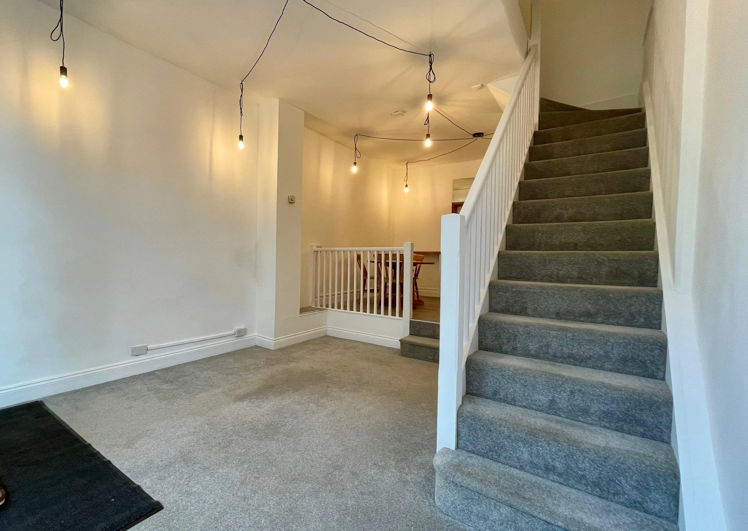An empty living room with hanging light bulbs, light-colored walls, a staircase with gray carpet, and a small wooden dining table visible in the background.