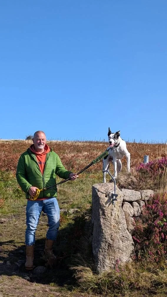A man holding a leash attached to a dog standing on a stone pedestal outdoors on a grassy hill, with a clear blue sky in the background.