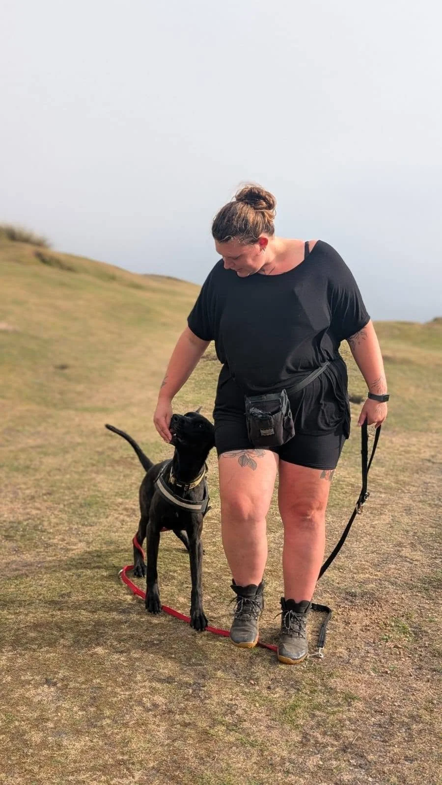A woman in black athletic clothing and hiking boots walks with a black dog on a leash across a grassy, hilly landscape during daytime.