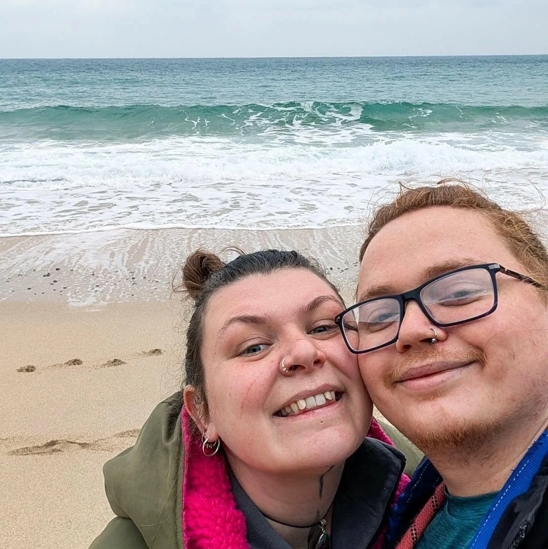 A smiling couple taking a selfie on a sandy beach with the ocean and waves in the background.