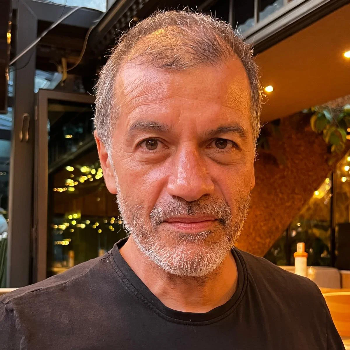 Close-up of a middle-aged man with gray hair and beard, wearing a black shirt, sitting in a warmly lit restaurant or cafe with decorative lighting and plants in the background.