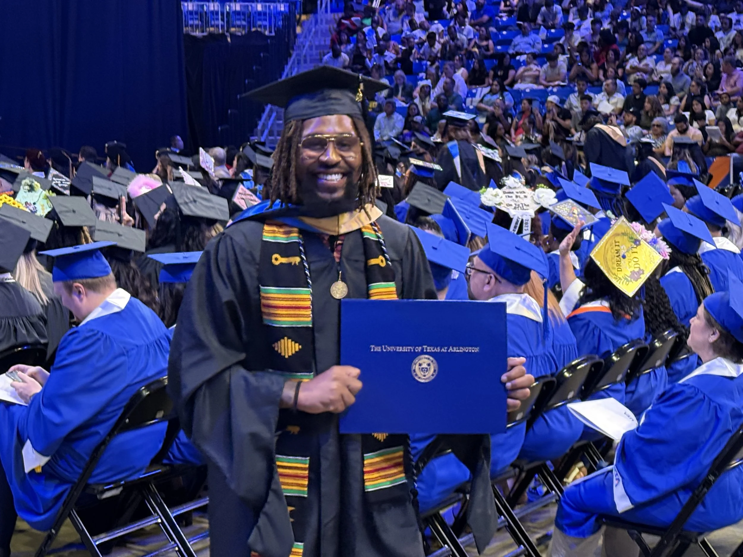 A man in graduation gown and cap holding a diploma from the University of Texas at Arlington, smiling at a graduation ceremony with many seated graduates in caps and gowns in the background.