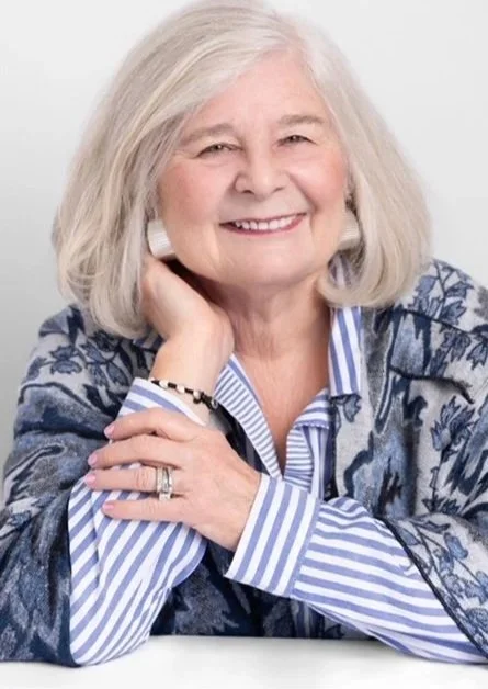 An elderly woman with shoulder-length gray hair, smiling, wearing a blue patterned blazer over a striped shirt, and posing with her hand resting on her chin.