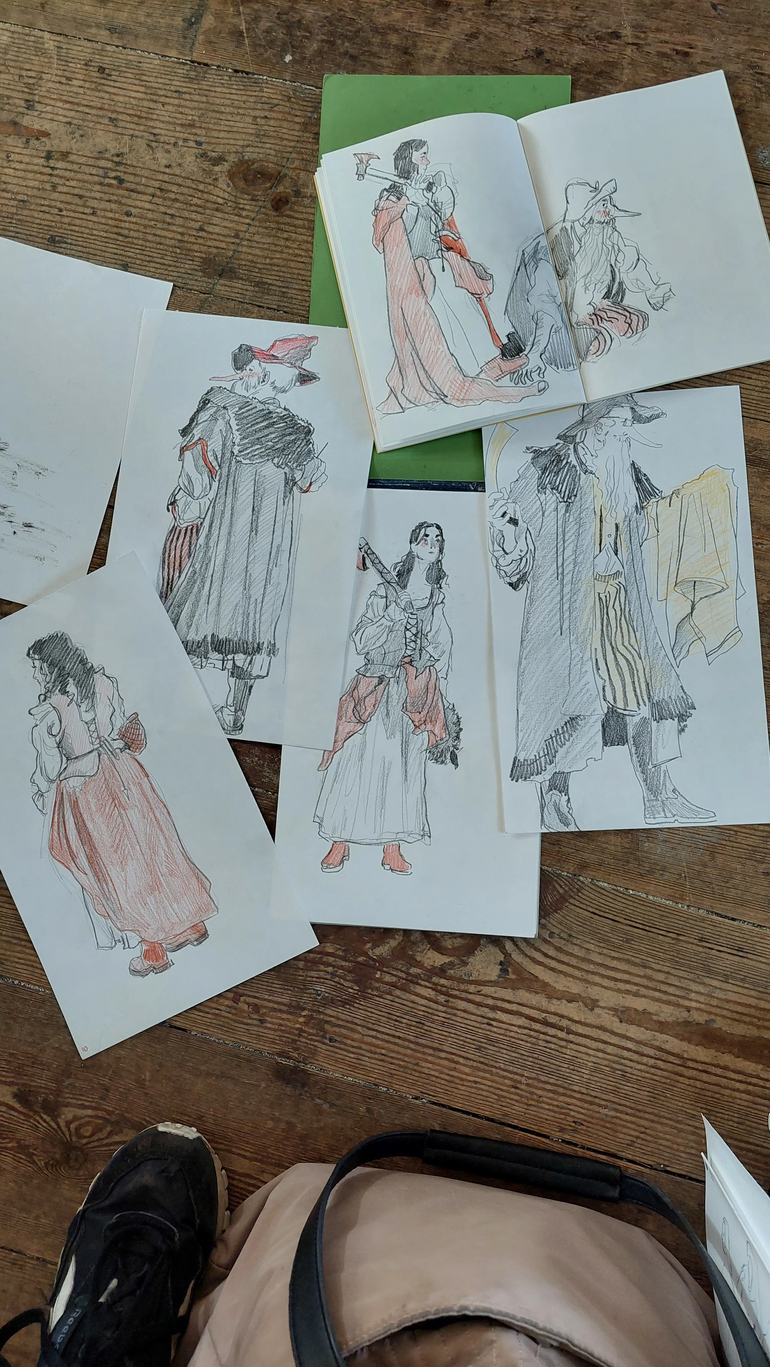 Several sketches of people in historical or fantasy costumes spread out on a wooden floor, with a person's foot visible at the bottom of the image.