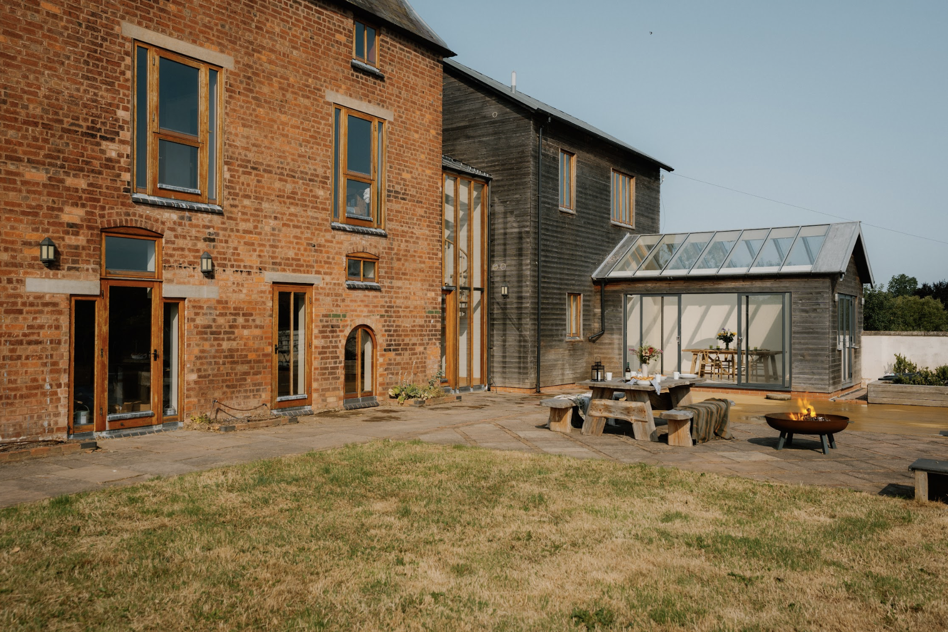 A modern brick and wooden house with a small outdoor patio, picnic table, fire pit, and a manicured lawn under a clear sky.