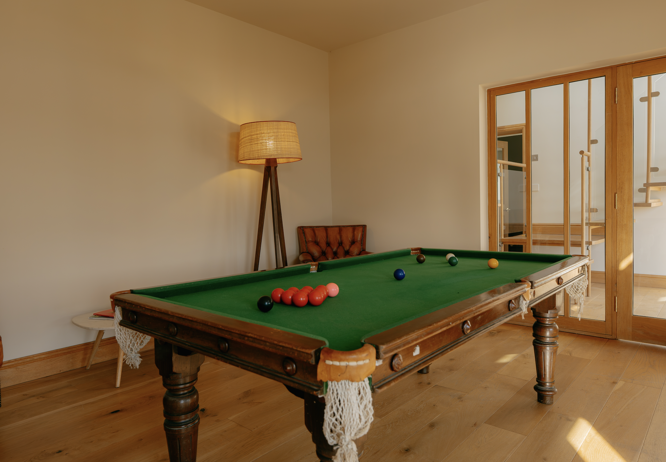 A wooden pool table with green felt, balls racked at one end, and balls scattered on the table. A standing lamp and a brown leather chair are visible in a room with light-colored walls and wooden flooring. A doorway with glass panels reveals a stairc
