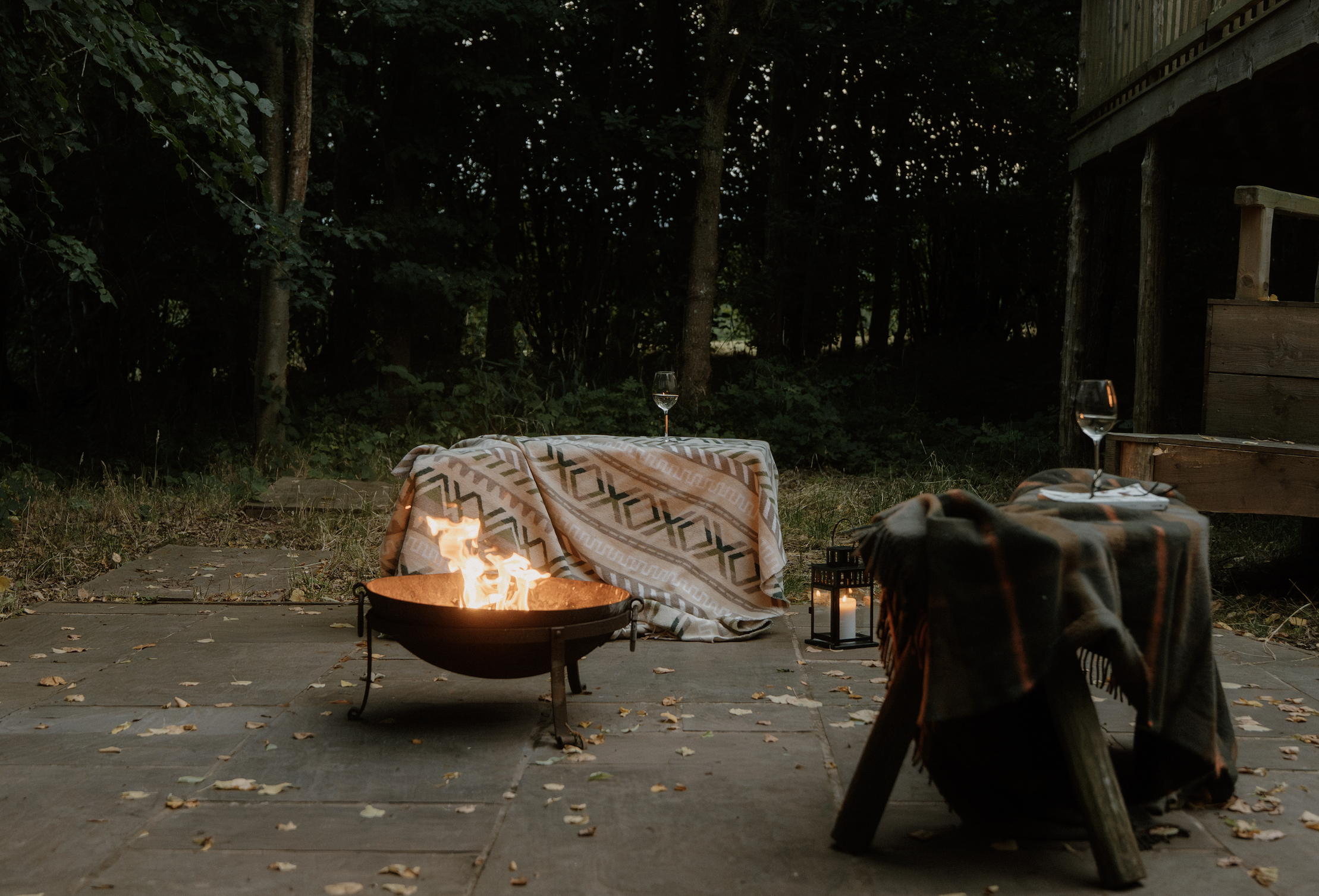 A cozy outdoor evening scene with a fire pit, blankets, wine glasses, candles, and lanterns set on a stone patio, surrounded by trees and a wooden deck.