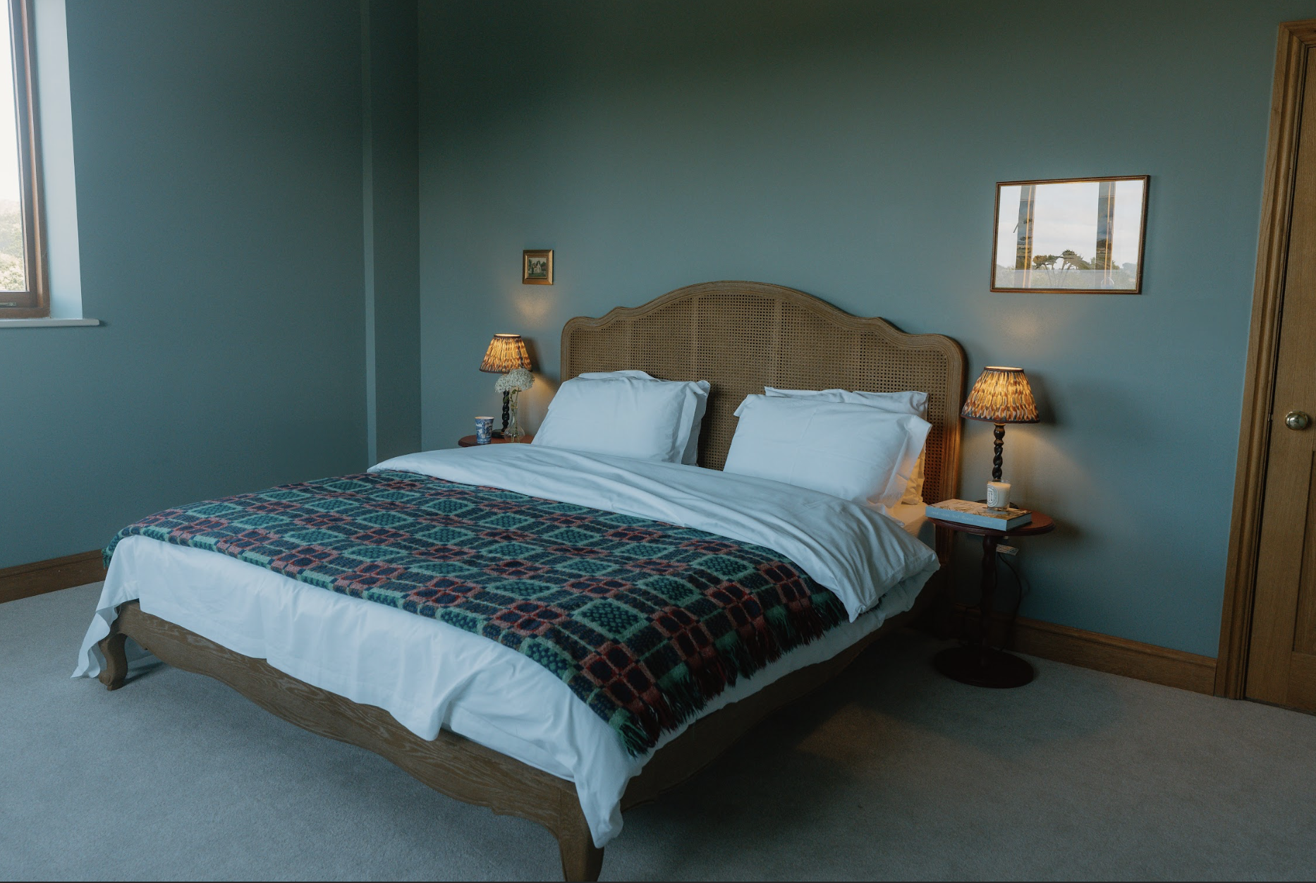 A neatly made bed with white pillows and a colorful patterned blanket in a cozy bedroom with blue walls, nightstands with lamps, and framed pictures hanging on the wall.