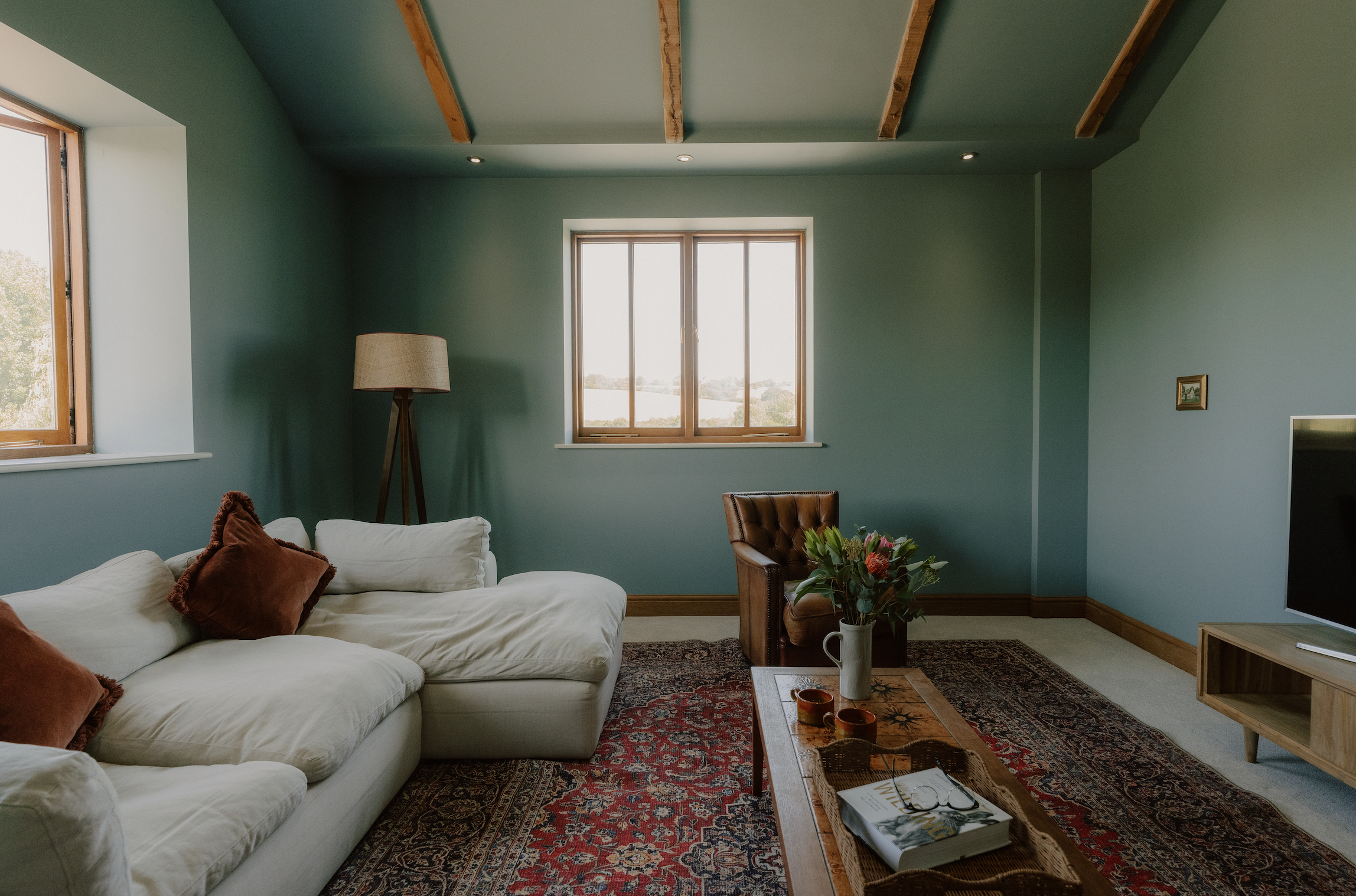 Living room with green walls, two windows, a white sofa with rust-colored pillows, a wooden coffee table with a book, candles, and a tray, an armchair, a carpet, a floor lamp, and a small picture on the wall.