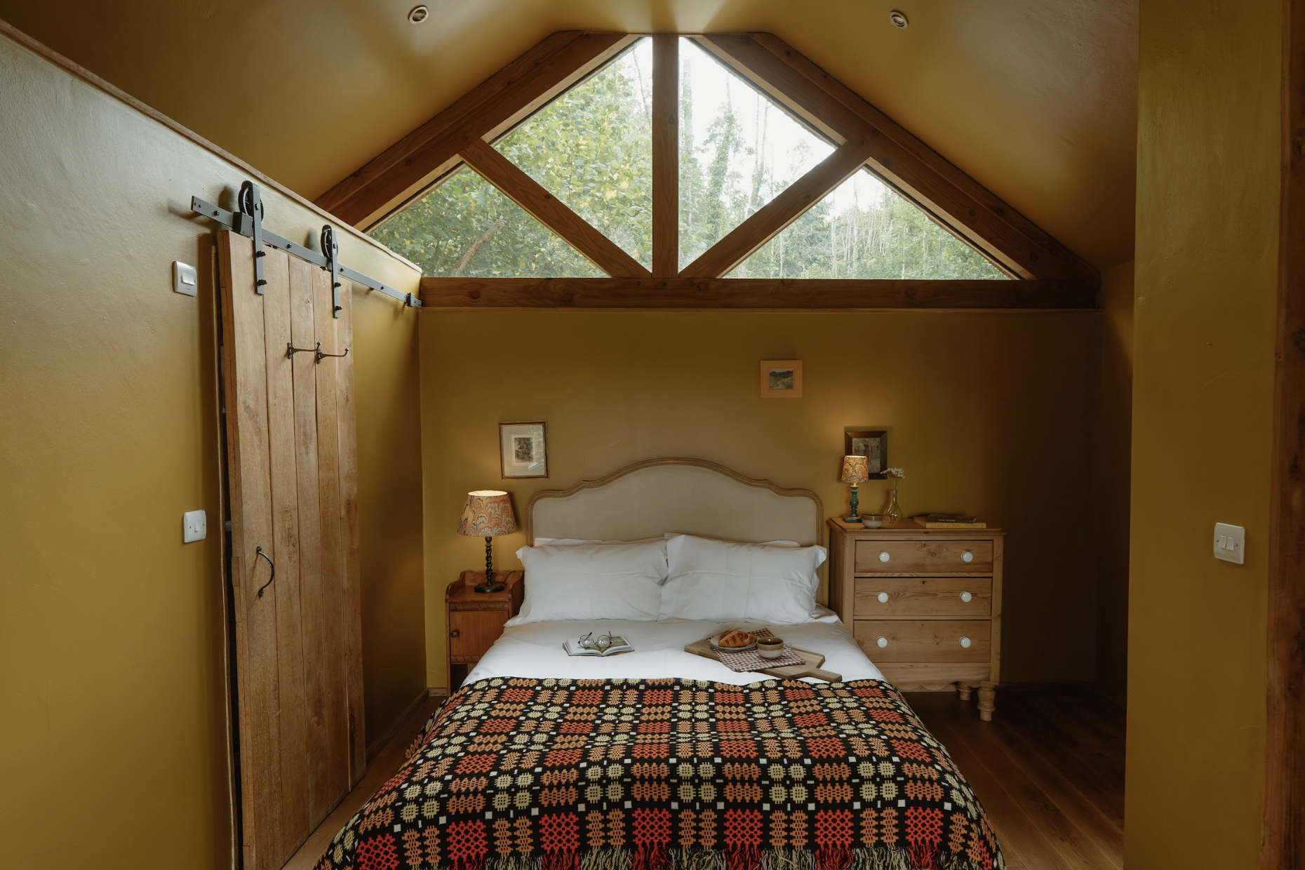 A cozy bedroom with yellow walls, a sloped ceiling, and a large triangular window showing green trees outside. The bed has white pillows, and a black, red, and beige patterned blanket. There are two wooden nightstands with lamps and decor, and a slid