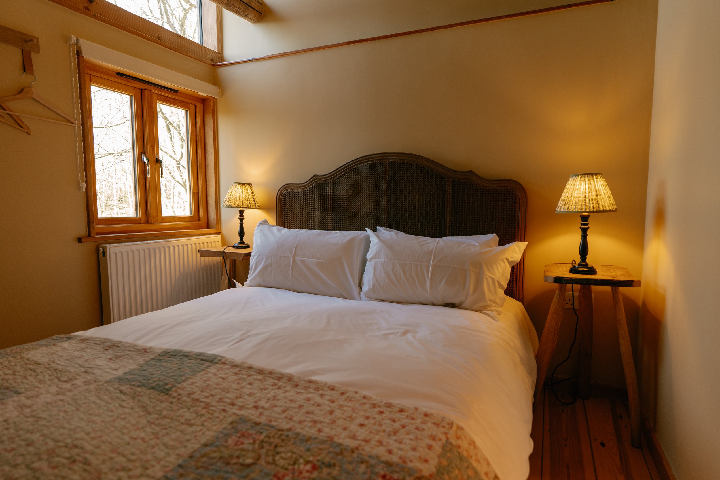 Cozy bedroom with a king bed featuring white linens, a wooden headboard, two bedside lamps, a window with wooden frames, and a quilted blanket.