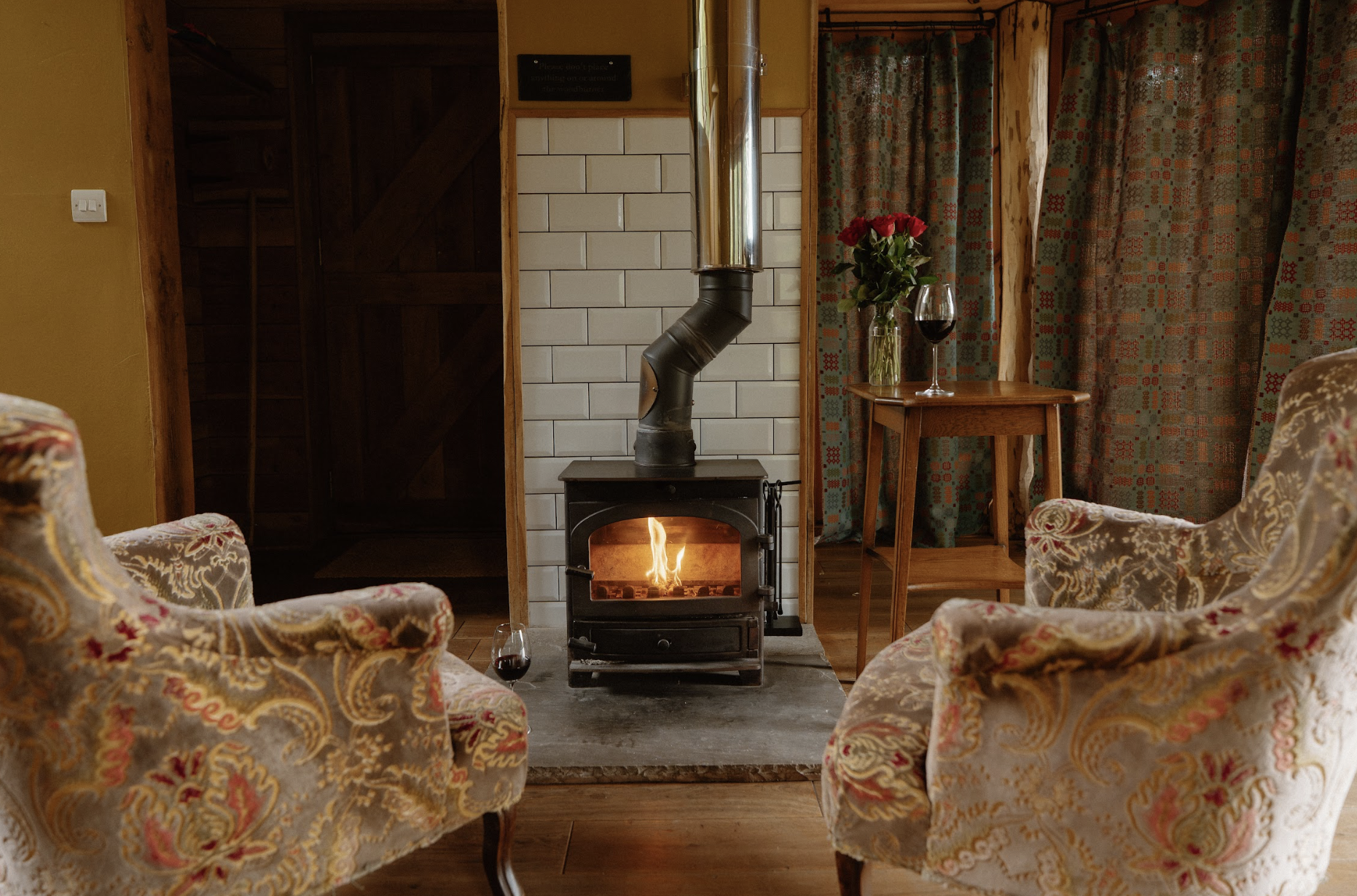 Cozy living room with two vintage armchairs, a wood-burning stove with a fire, a table with a vase of red roses and a glass of red wine, wood and patterned curtains, and warm wooden accents.