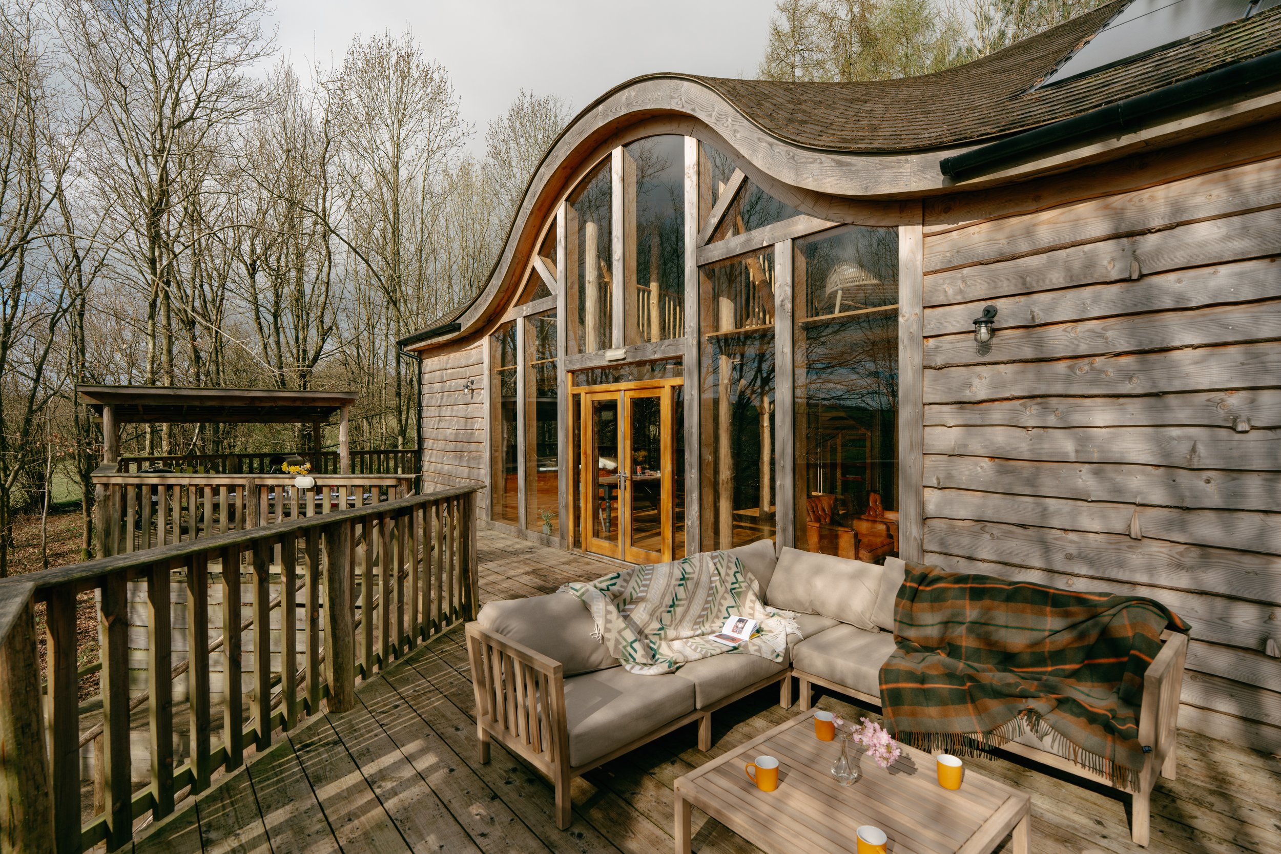 Wooden cabin with large glass windows, a curved roof, and an outdoor deck with seating and blankets, surrounded by trees.