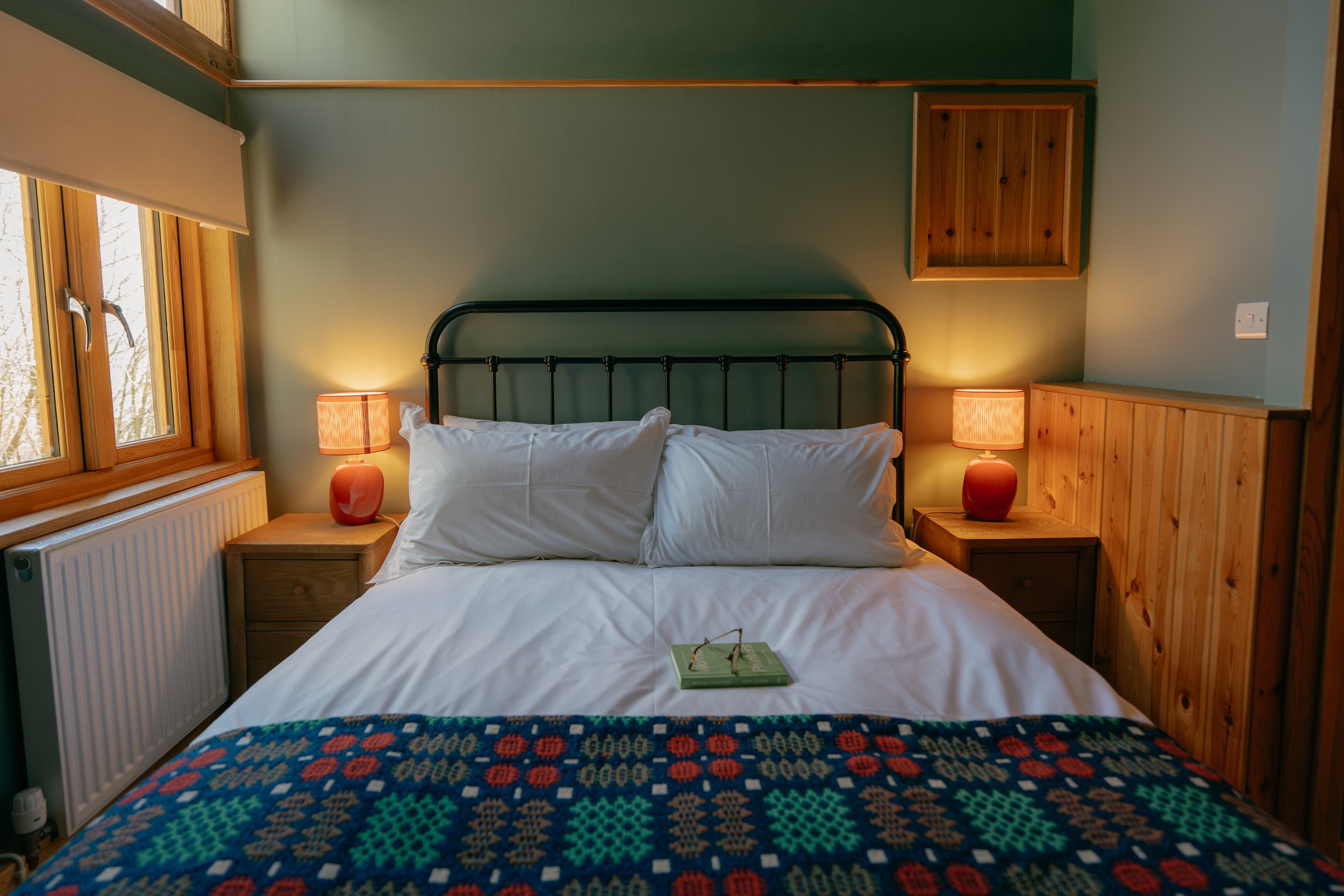 Cosy bedroom with a double bed, white bed linen, decorative blanket, wooden side tables, red lamps, green painted walls, and a window with a view.