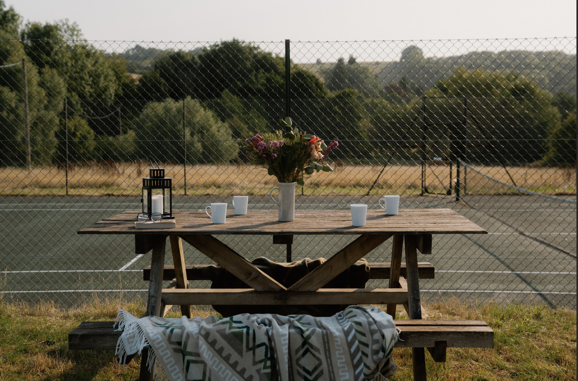 A picnic table with a vase of flowers, four white cups, and a lantern, set outdoors in front of a tennis court and a chain-link fence, with trees and open land in the background.