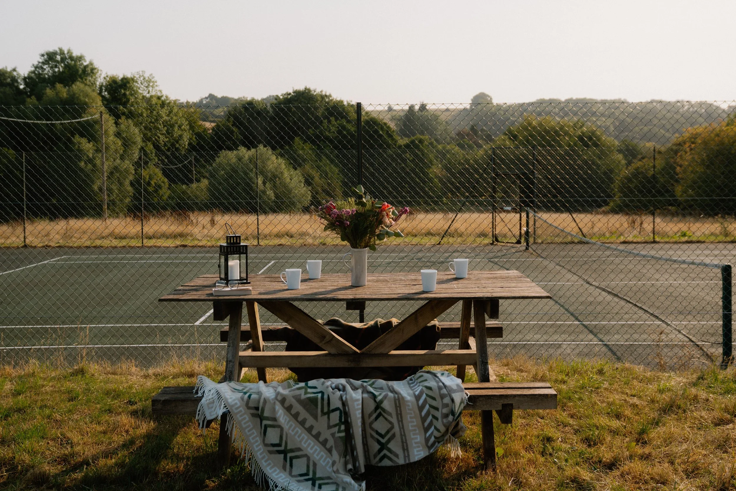 A wooden picnic table with a floral centerpiece and white cups on a tennis court, enclosed by a chain-link fence, with trees and hills in the background during daytime.