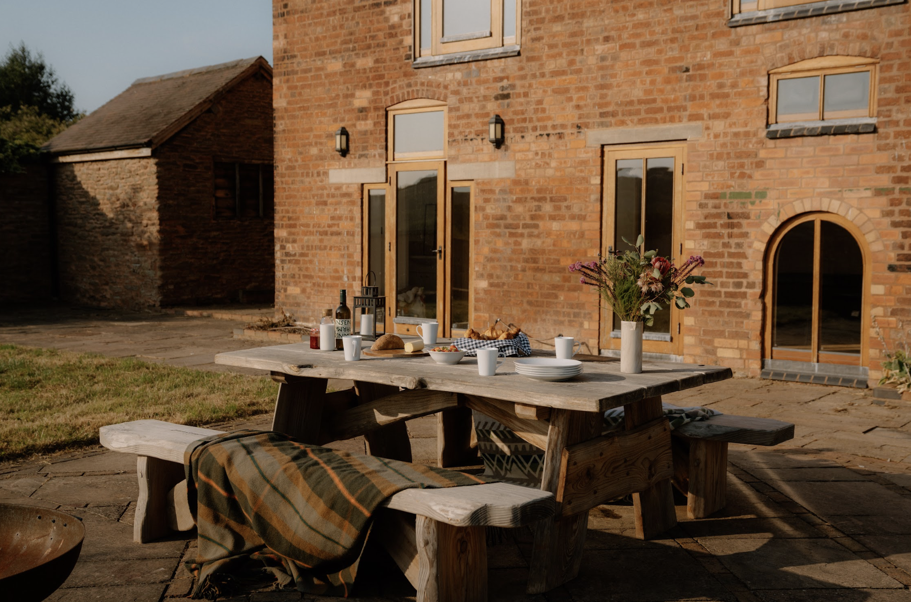 Outdoor dining table set with plates, cups, bread, and a flower vase in front of a brick house.