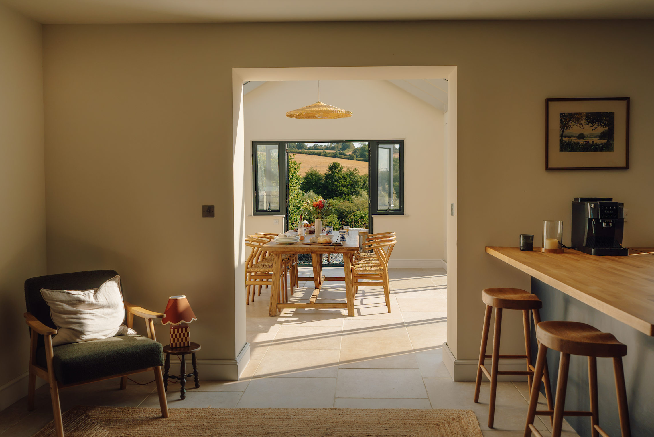 A dining area seen through a doorway in a home, with a wooden dining table, chairs, sunlight, open windows revealing a landscape of trees and fields, and a hanging light fixture.