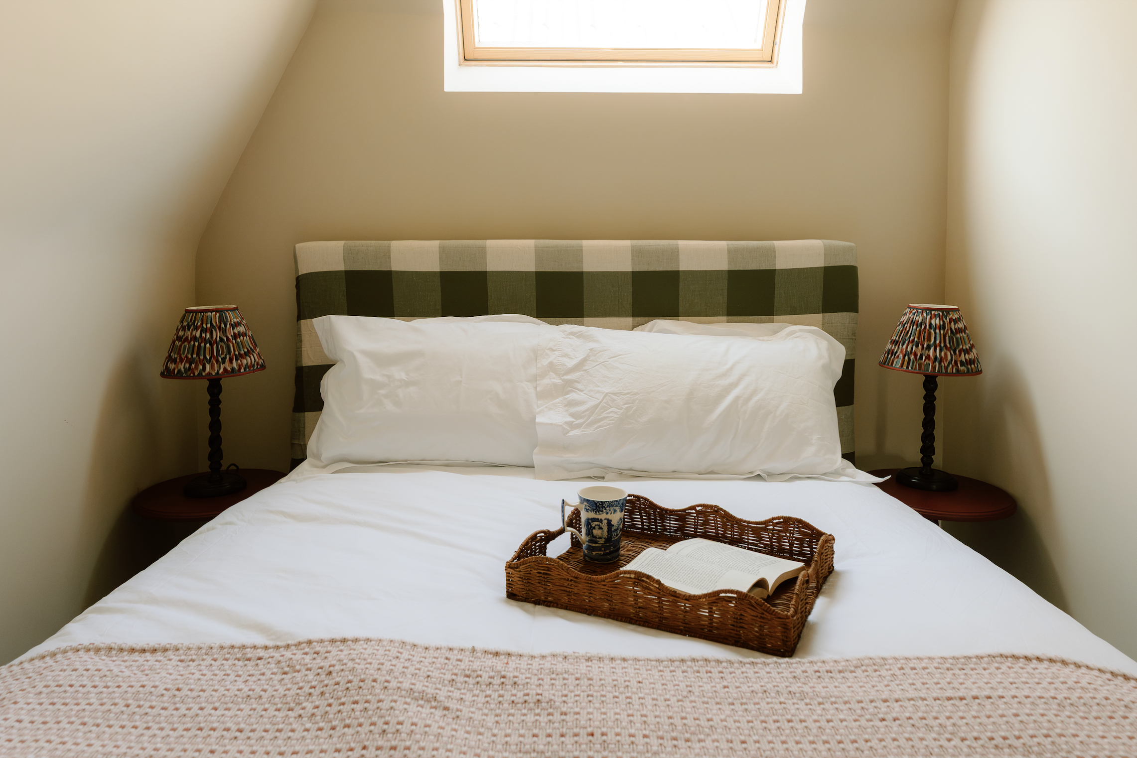 A cozy bedroom with a bed and two bedside lamps with colorful patterned shades, a window above the bed, and a tray with a cup and an open book on the bed.