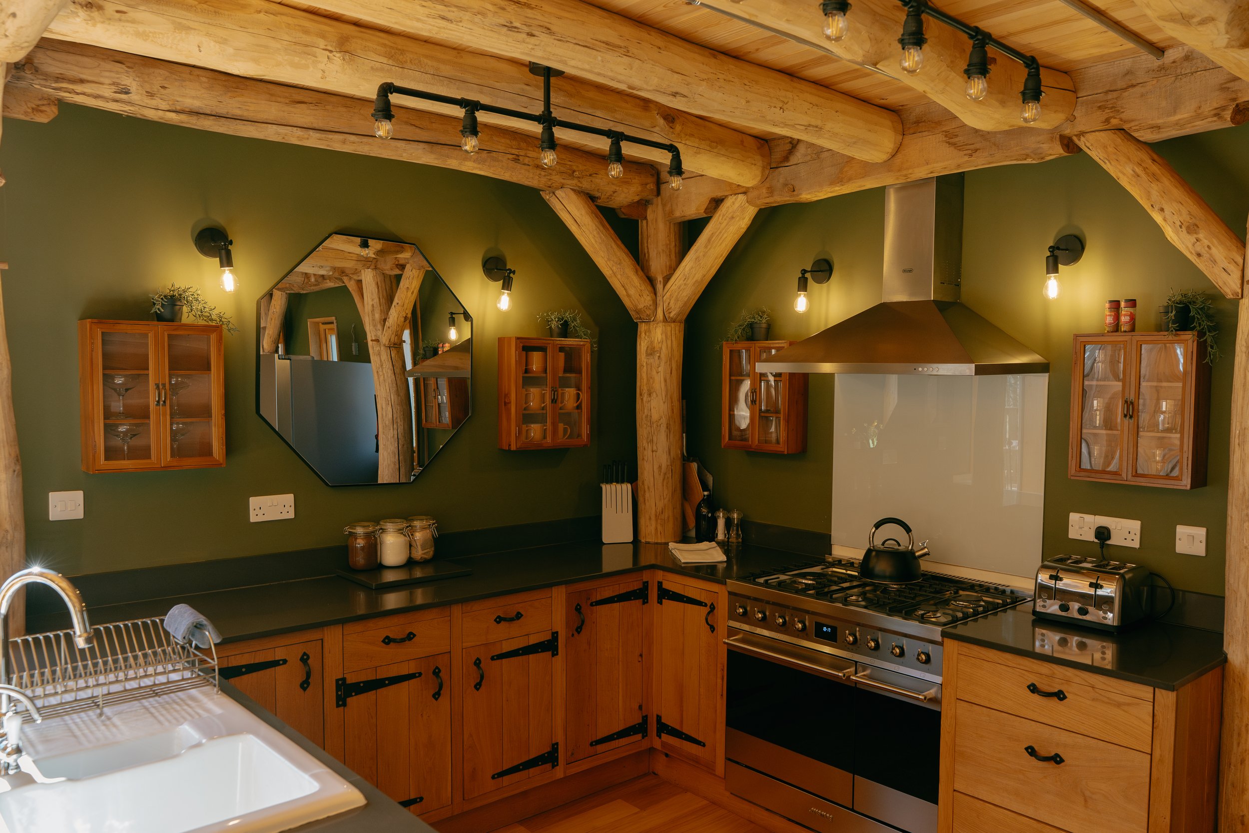Rustic kitchen with wooden beams, green walls, and modern appliances, including a stove and toaster. Features wooden cabinets, a sink, and a geometric mirror.