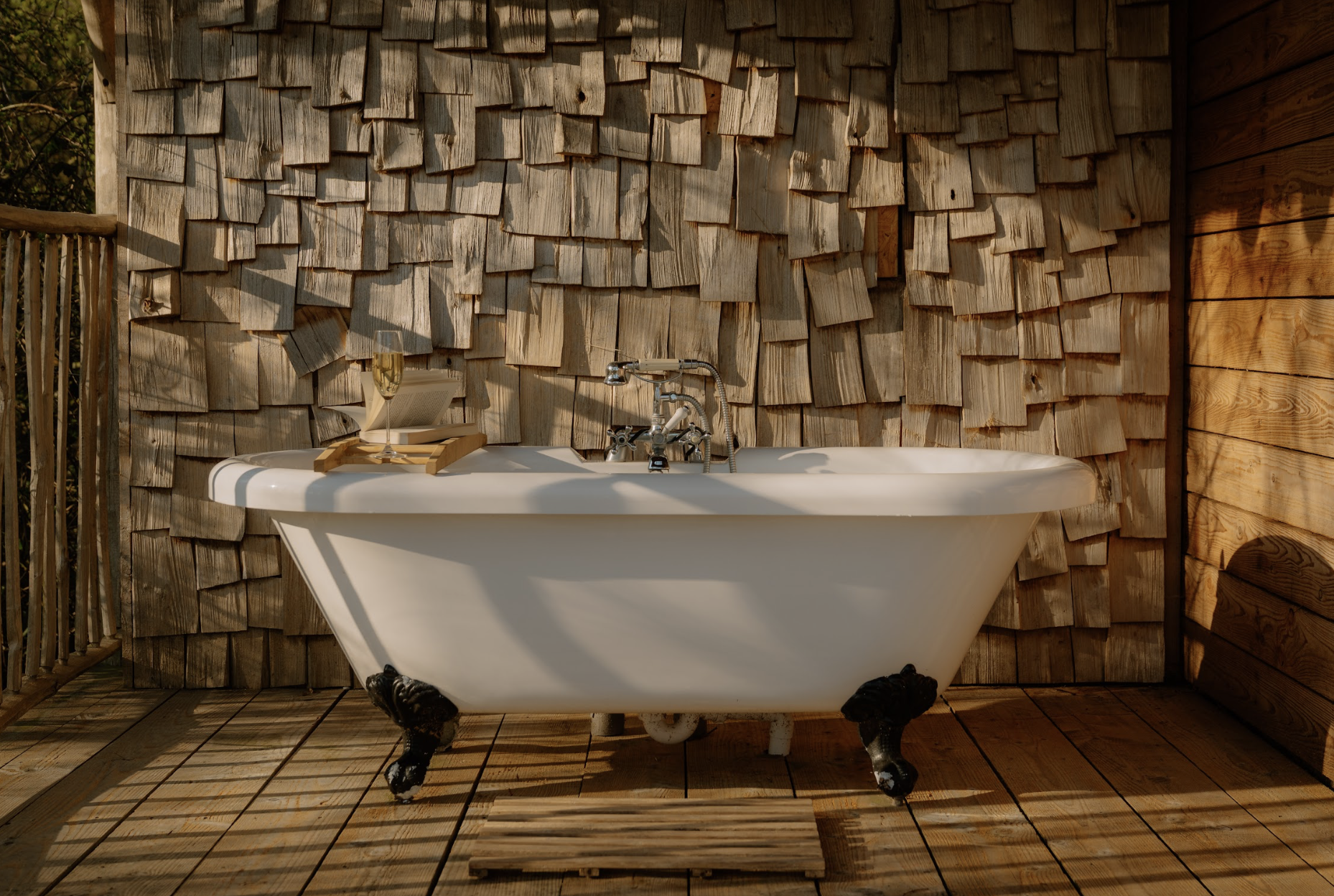 Outdoor bathtub with clawfoot design, placed on a wooden deck with wooden walls in the background, and a glass of champagne and an open book on a tray on the edge of the bathtub.
