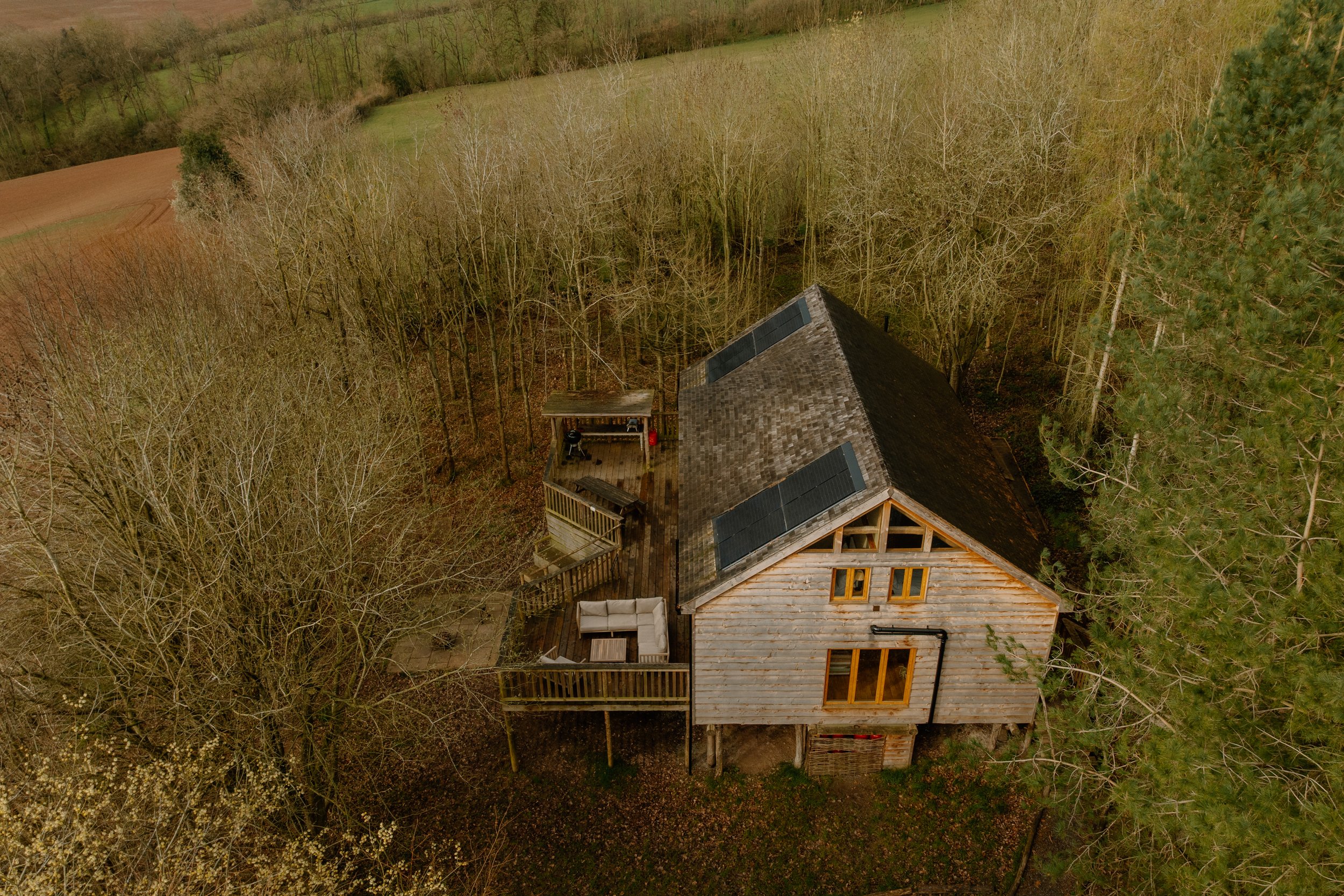 Aerial view of a secluded wooden cabin with solar panels on the roof, surrounded by leafless trees and open fields.