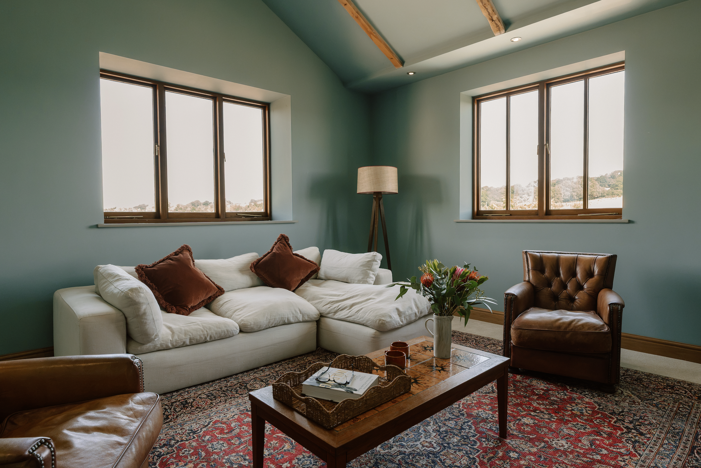 A cozy living room with a white sectional sofa with brown pillows, a wooden coffee table with a basket and books, a leather armchair, a patterned rug, tall windows with wooden frames, a floor lamp, and green painted walls with exposed wooden beams ce