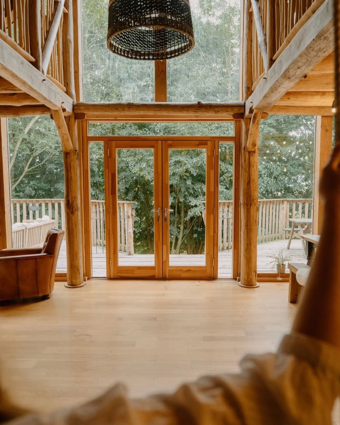 A spacious interior with wooden walls, ceiling, and floor, featuring large glass doors leading to an outdoor deck with a picnic table and surrounding trees.
