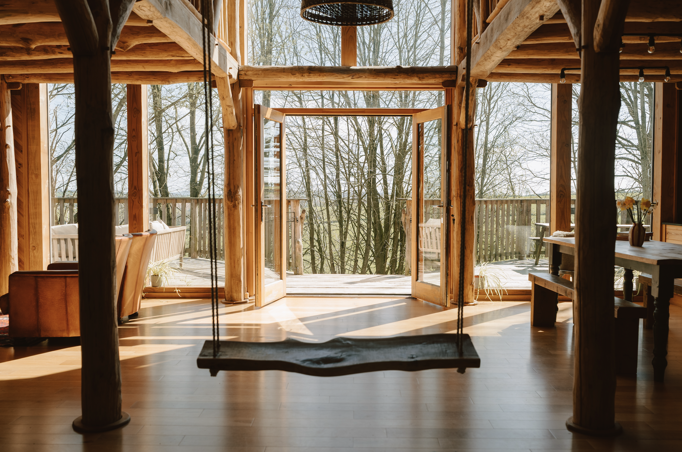 Sunlit double-height entrance inside Journeyman Treehouse with open doors to the woodland deck and a suspended timber swing