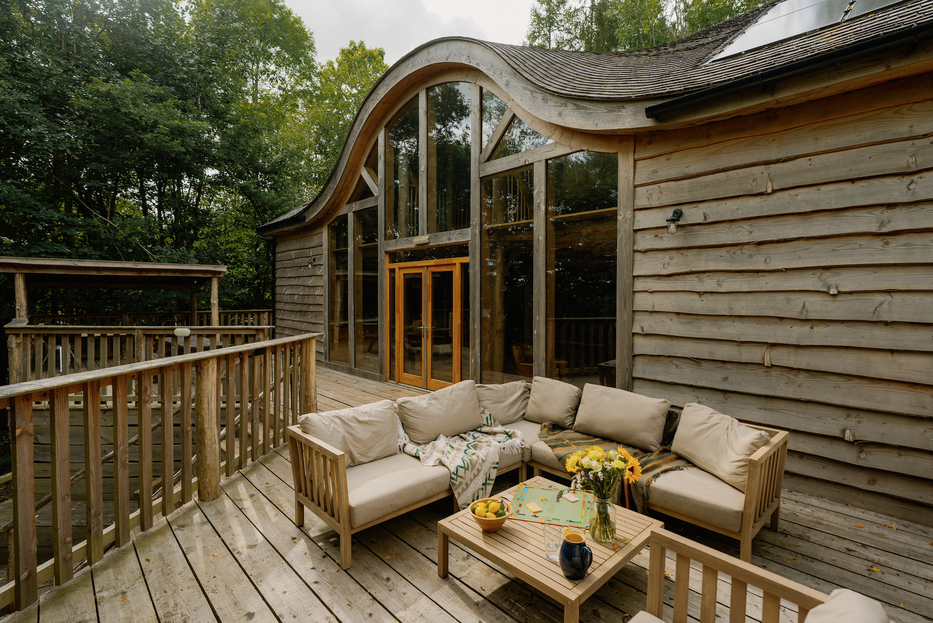 A wooden deck with outdoor seating, including a beige cushioned sectional sofa with pillows, a coffee table with flowers, a bowl of apples, and beverages. The house behind has curved glass windows and a wooden exterior surrounded by green trees.