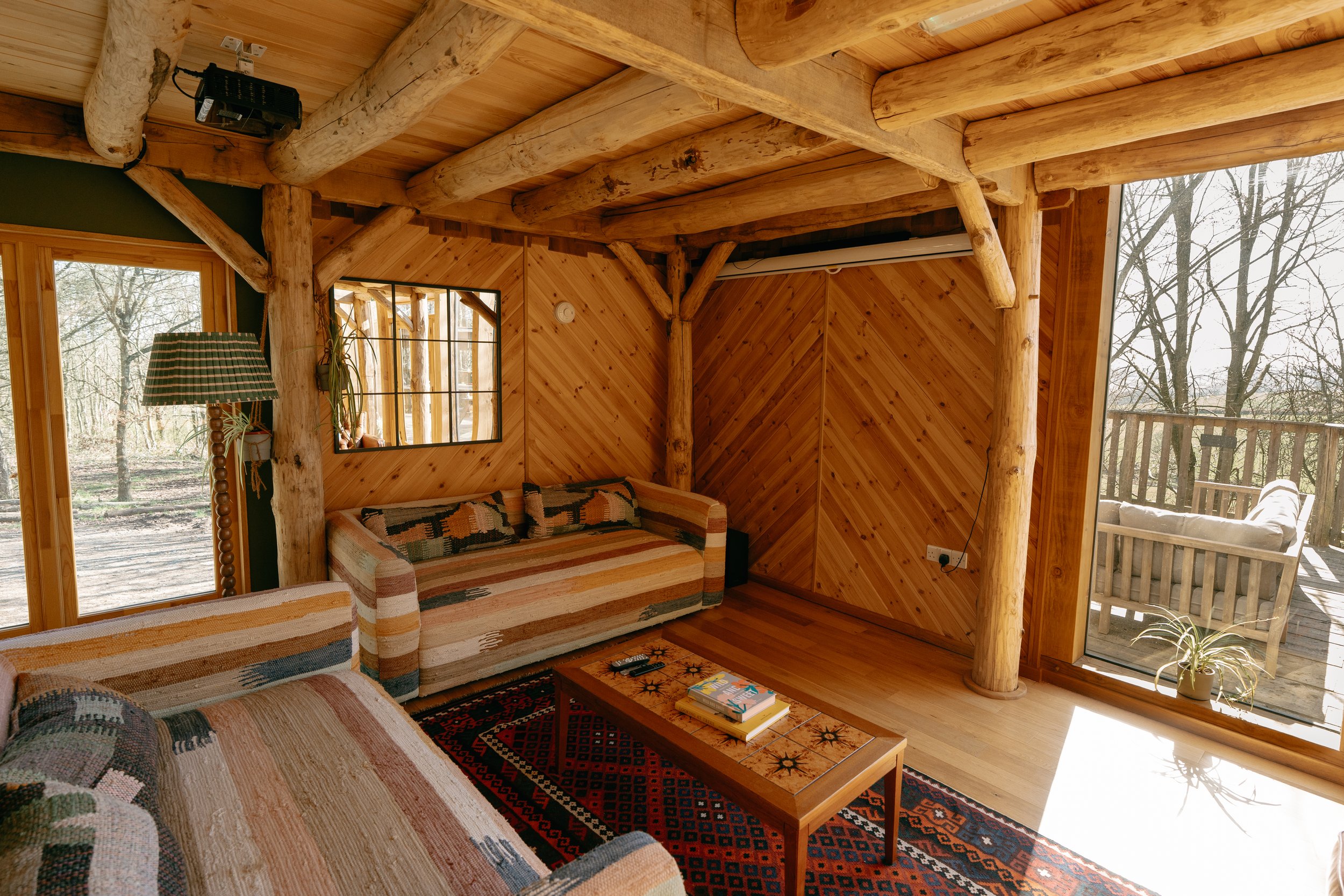 Cosy log cabin living room with wooden walls, sofas with striped fabric, a coffee table with books, and a view of an outdoor deck with trees.