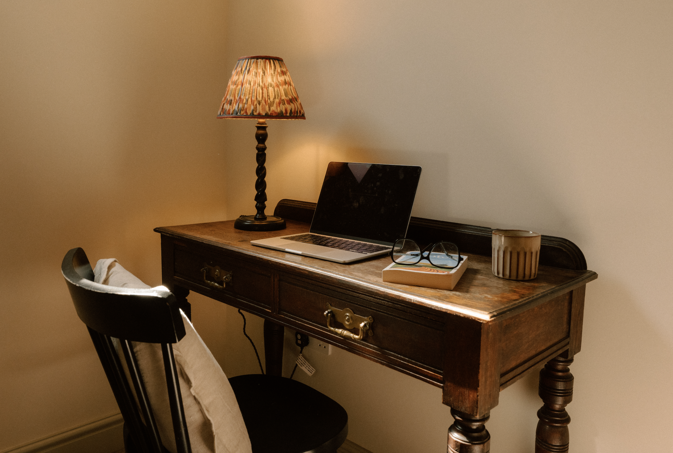 A wooden desk with a lamp, a laptop, a pair of glasses, a book, and a coffee mug, against a beige wall.