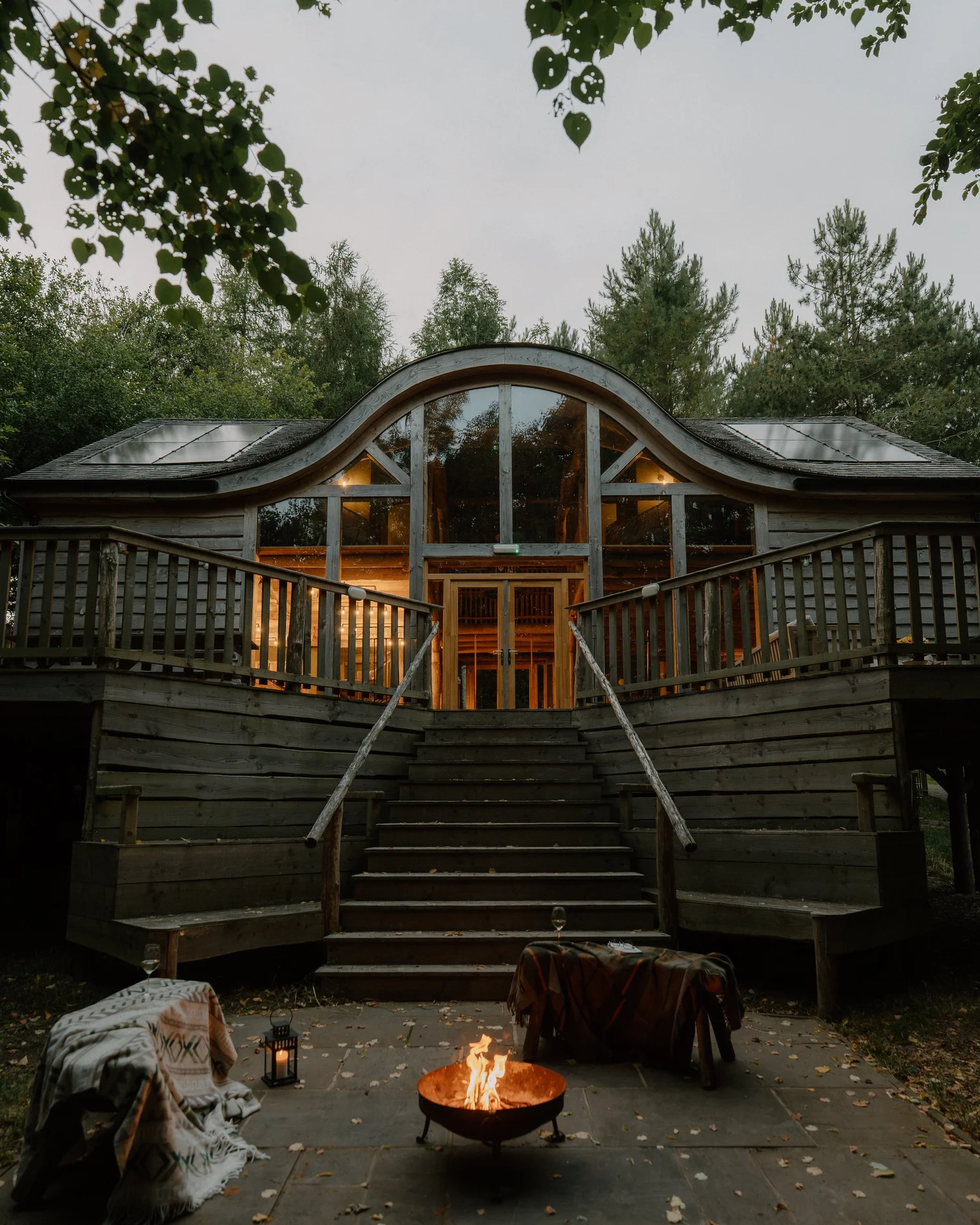 Front view of Journeyman Treehouse at dusk with its curved timber roof, woodland deck and glowing fire pit in Herefordshire