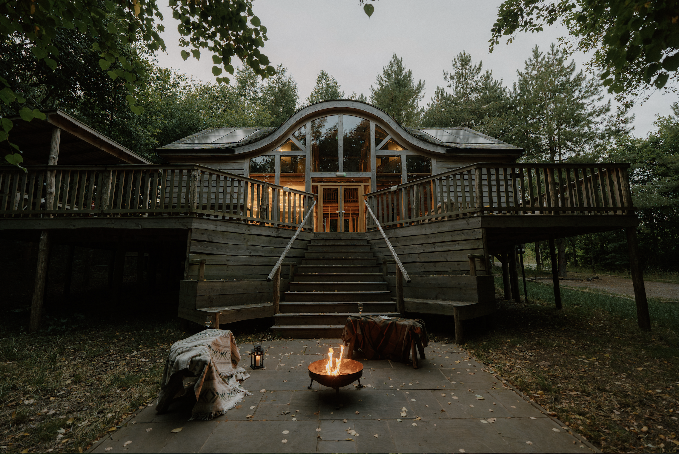 A wooden house with a large arched window and balcony, surrounded by trees, with a fire pit and seating area outdoors, during dusk or evening.