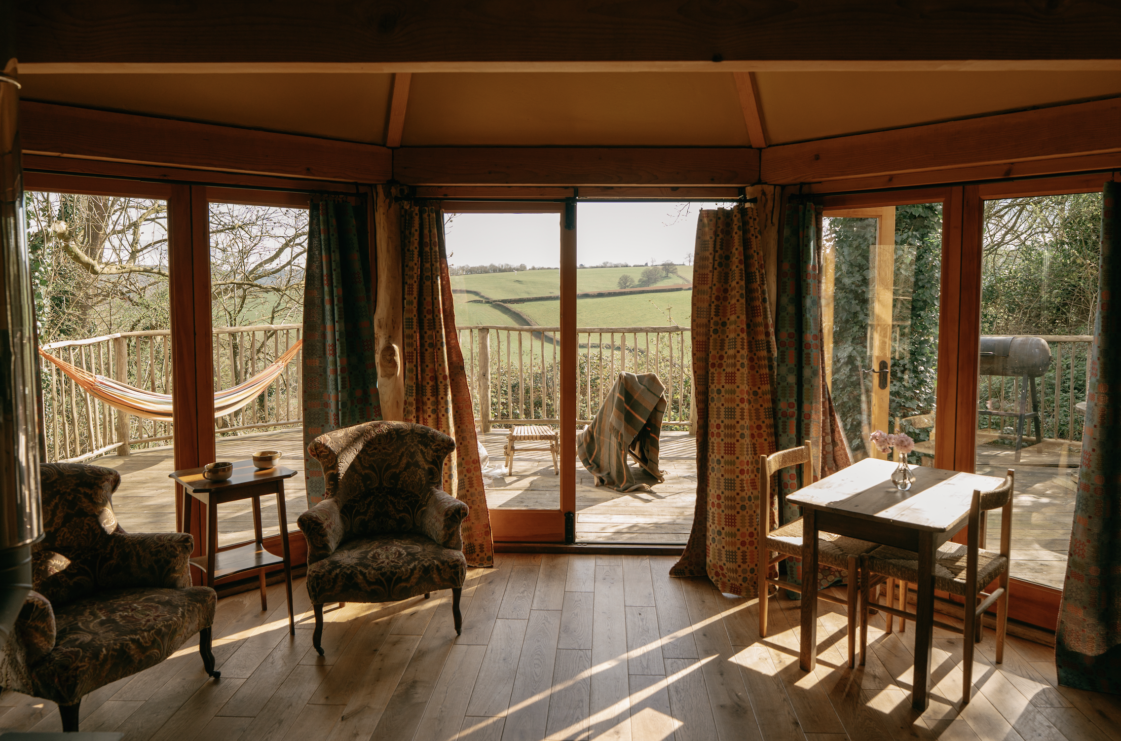 Living room with large glass sliding doors opening to a wooden deck with a hammock, a chair with a towel draped on it, and a scenic view of green hills and trees.