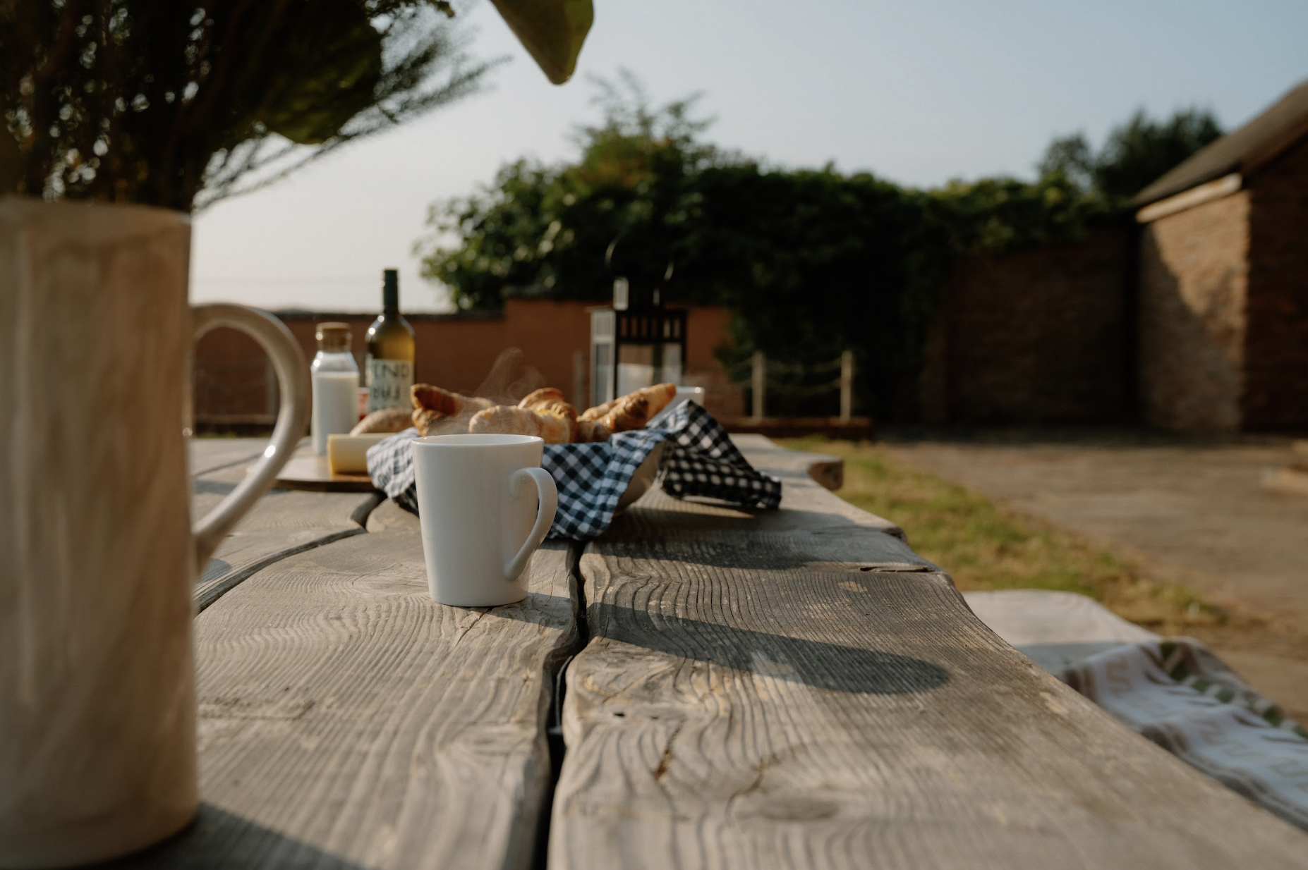 An outdoor wooden table set for breakfast with white ceramic mugs, a basket of croissants with steam, a bottle of wine, and a small bottle of milk, on a checkered cloth, with a blurred background of a brick wall, garden, and sky.