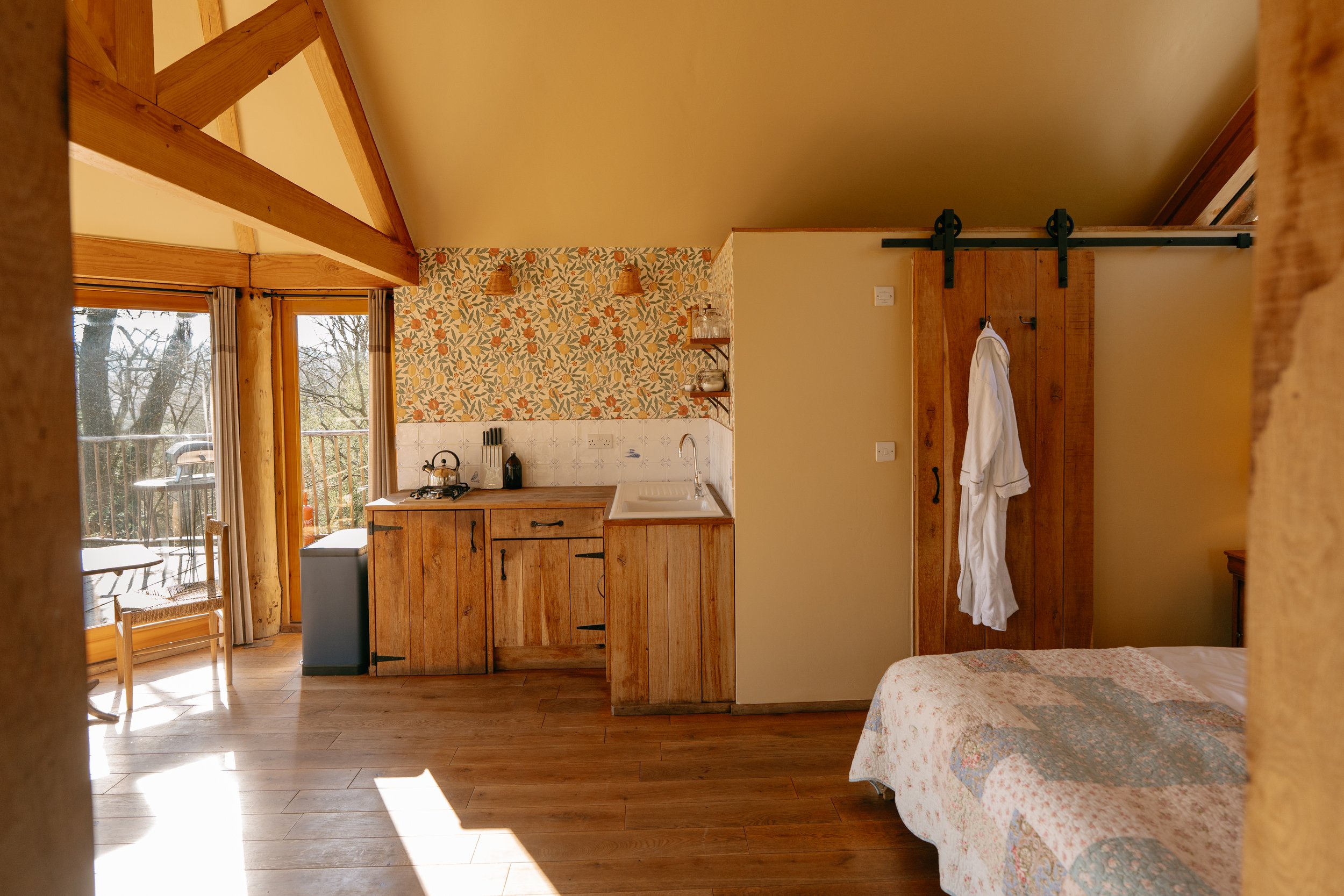 Cosy rustic cabin interior with wooden beams, a kitchenette, and a sliding barn door. There's floral wallpaper, a bed with a quilt, a robe hanging on the door, and an outdoor view through glass doors.