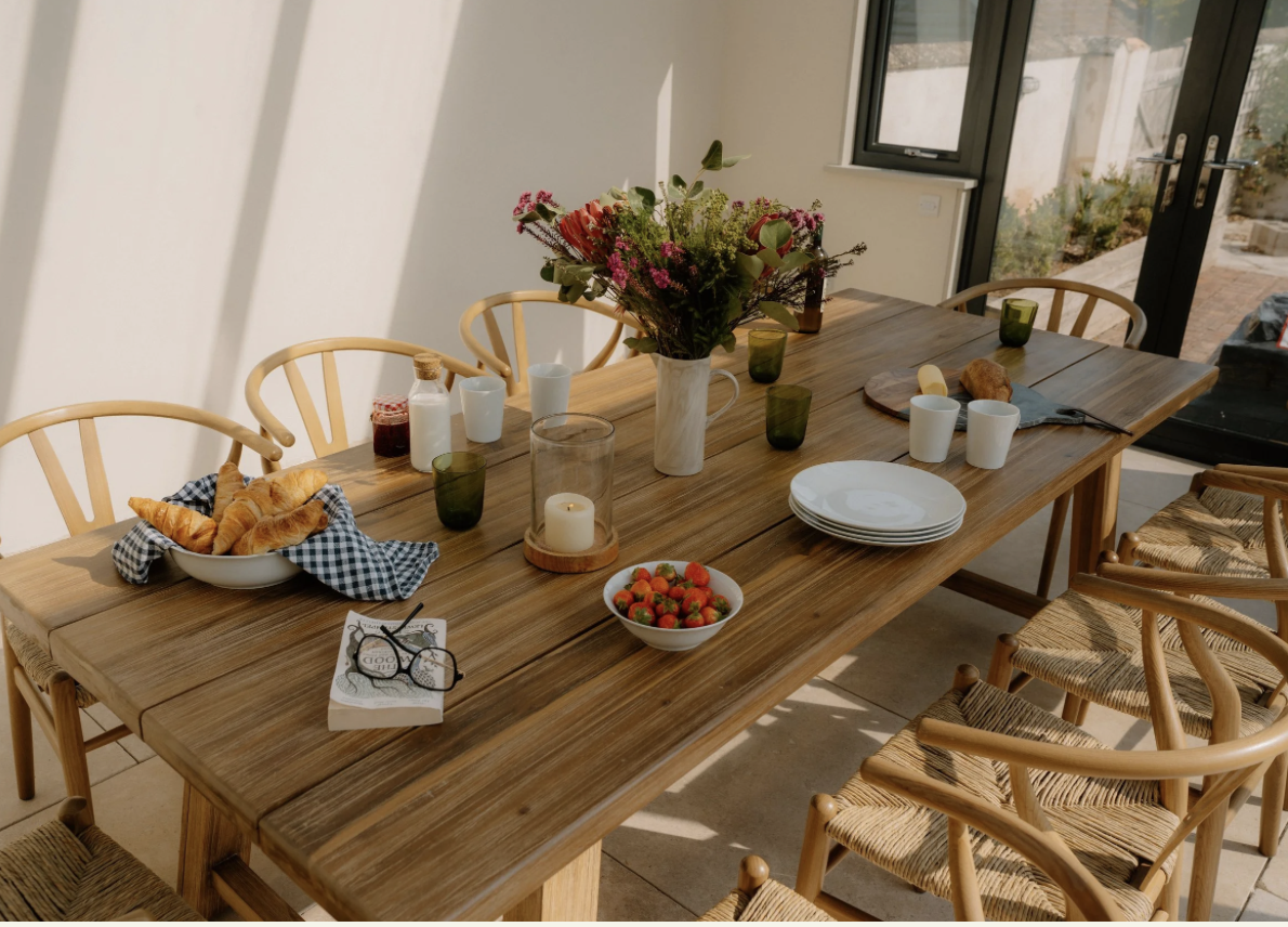 Wood dining table set with white plates, a bowl of strawberries, croissants with a checkered napkin, glass cups, green glasses, a candle, flowers in a white vase, and bread on a cutting board, in a sunlit room with a glass door.