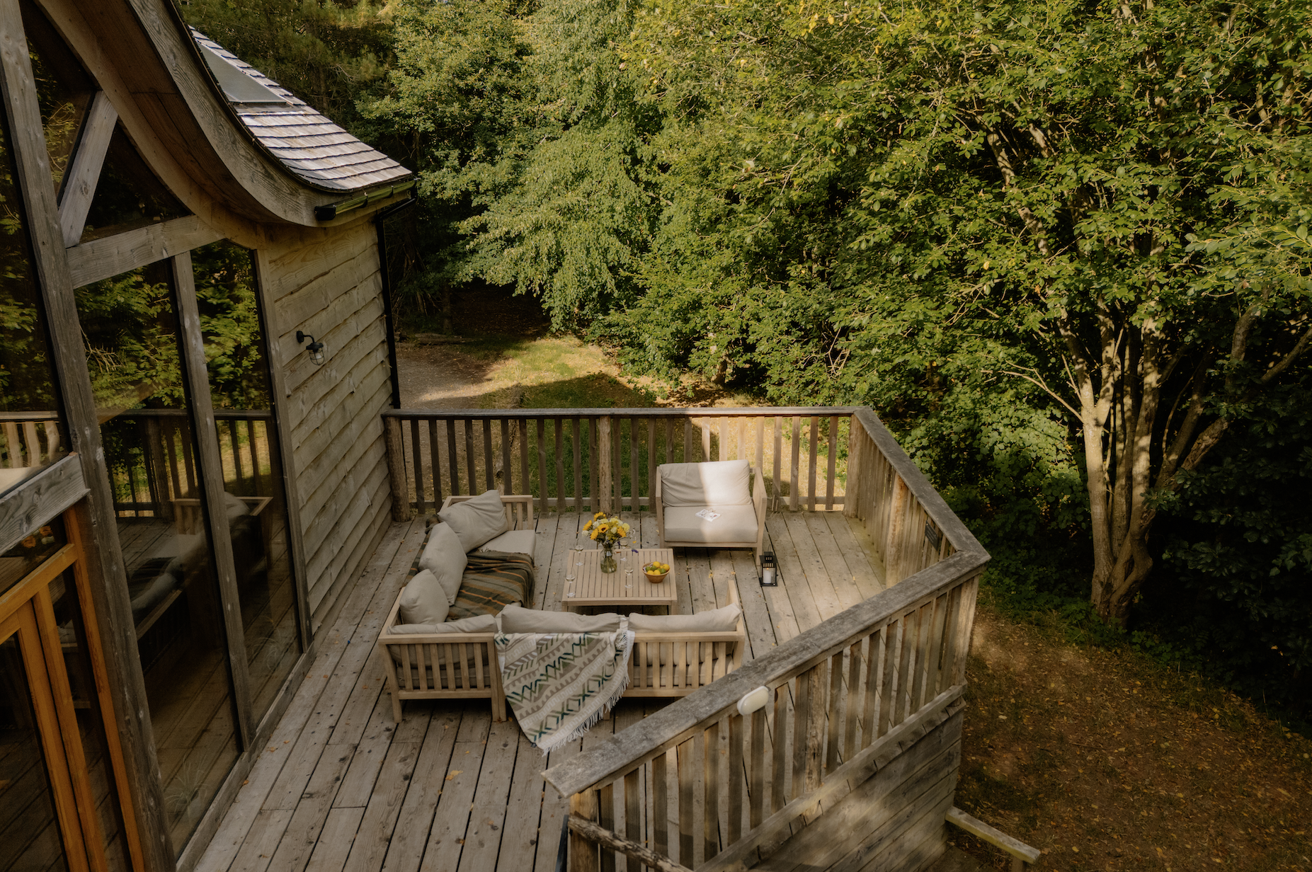 An outdoor wooden deck surrounded by lush green trees, furnished with a sectional sofa, an armchair, a coffee table with a vase of sunflowers and bowls of fruit, and a lantern.